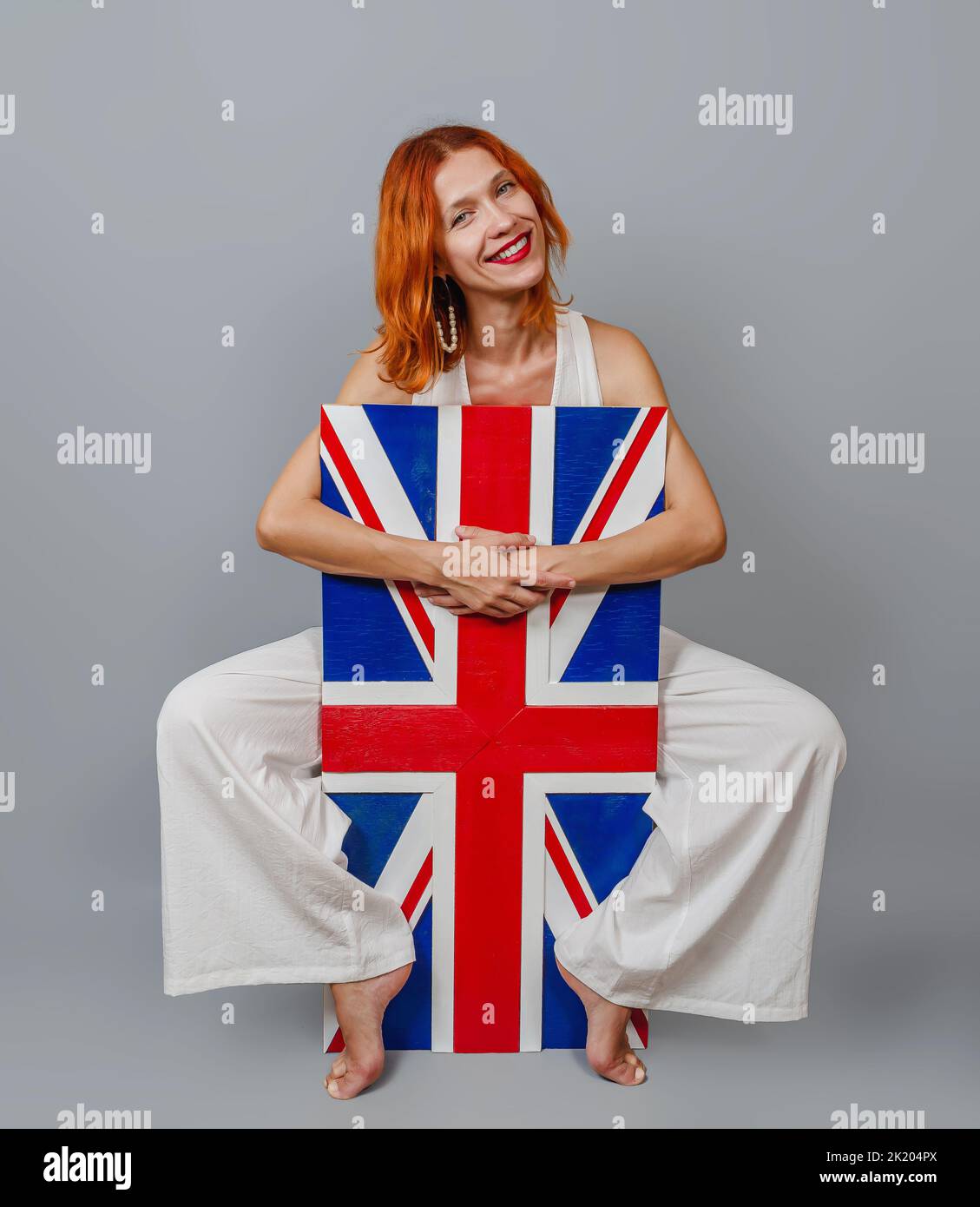 Donna dai capelli rossi dall'espressione allegra, in eleganti pantaloni larghi bianchi, abbraccia la bandiera britannica con le braccia e le gambe. Sfondo grigio. Foto Stock