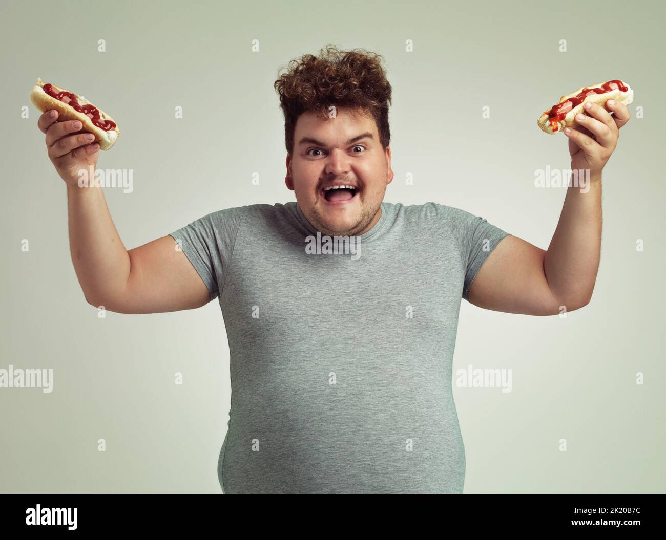 Più è meglio. Studio shot di un felice uomo sovrappeso in possesso di 2 hot dog in aria. Foto Stock