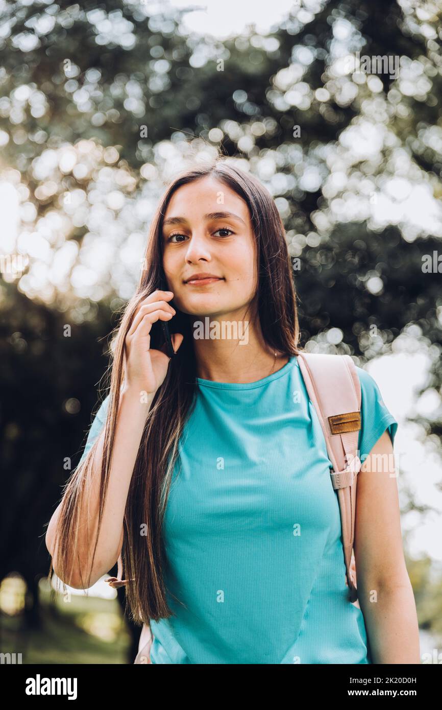 Ragazza studentesca adolescente che indossa una t-shirt acquamarina, facendo una telefonata alla sua famiglia nel parco. Retroilluminazione del sole Foto Stock
