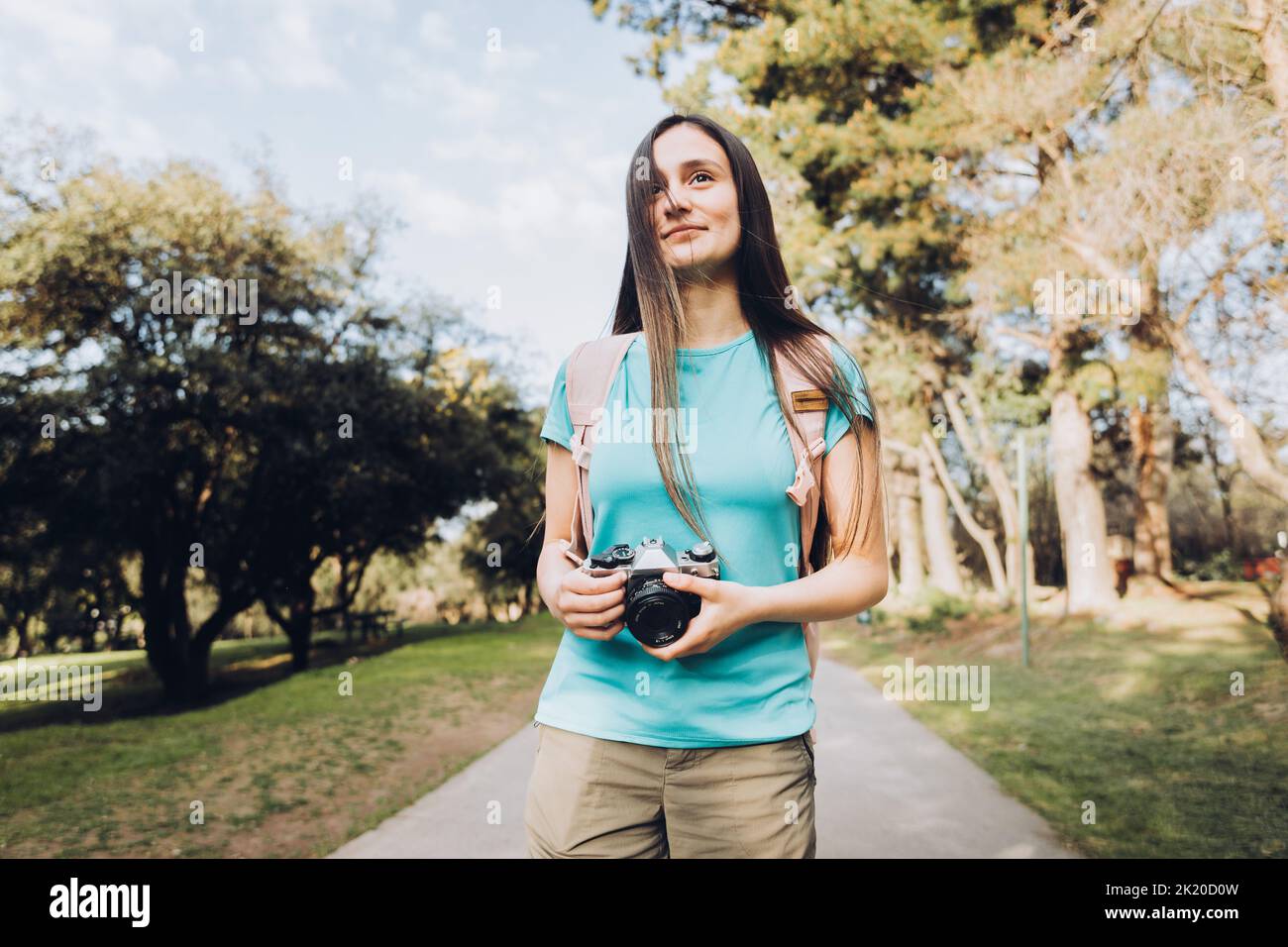 Giovane donna turistica che viaggia, indossando una t-shirt turchese e uno zaino, scattando foto mentre cammina nel parco. Foto Stock