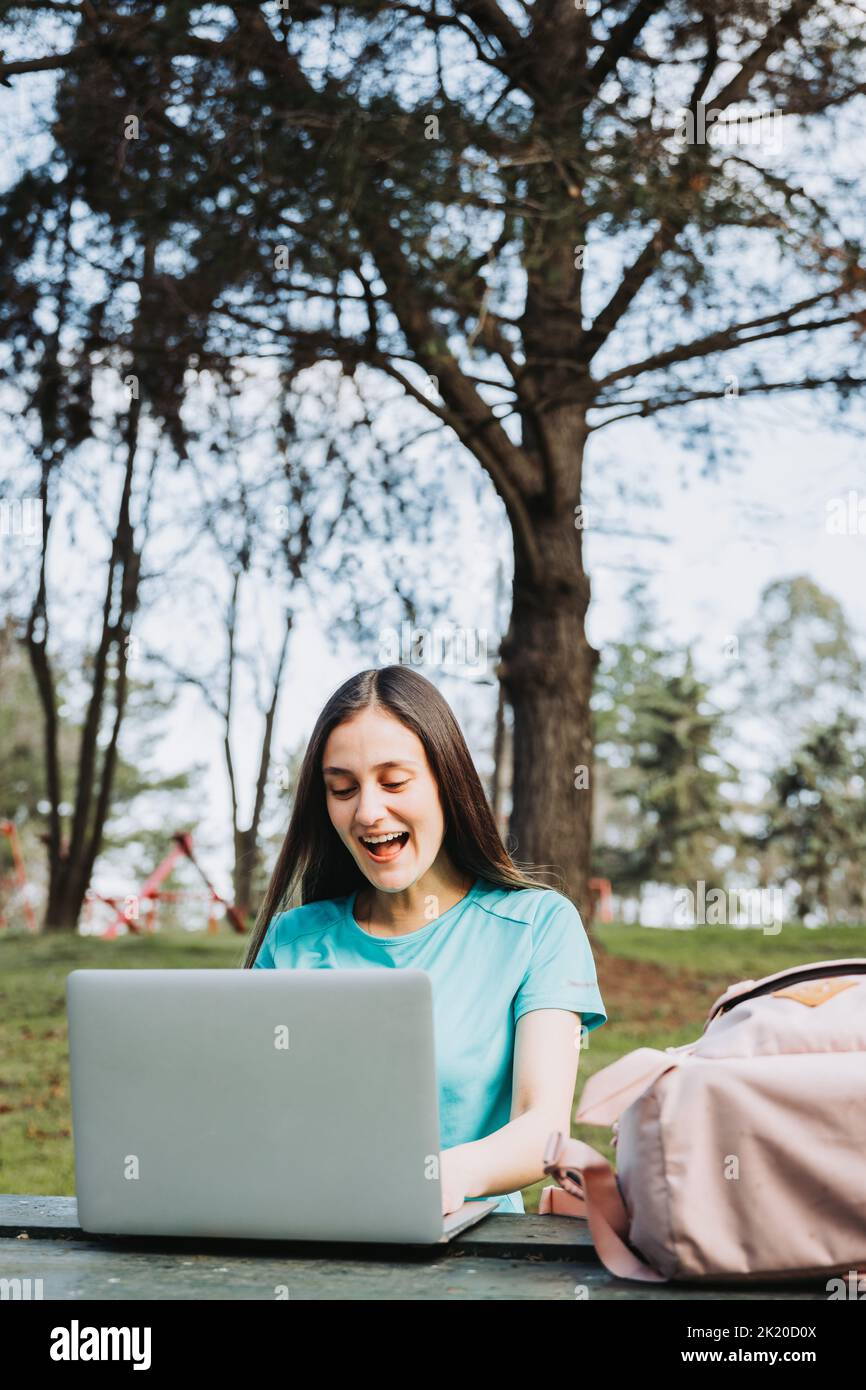Studentessa universitaria che ride e usa il suo computer portatile nel parco del campus universitario. Maglietta turchese Foto Stock