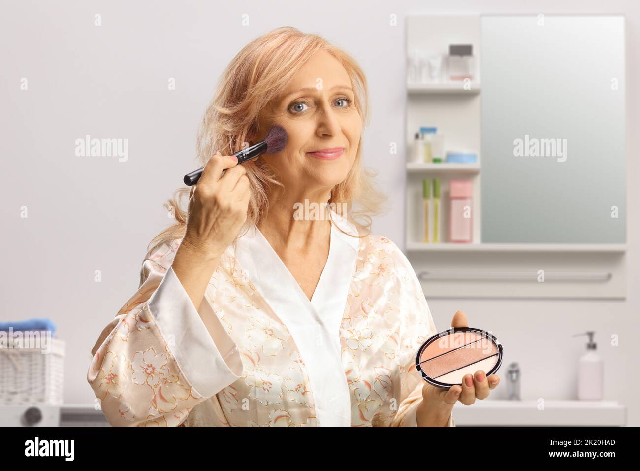 Donna matura in un accappatoio che applica il make up utilizzando una spazzola all'interno di un bagno Foto Stock