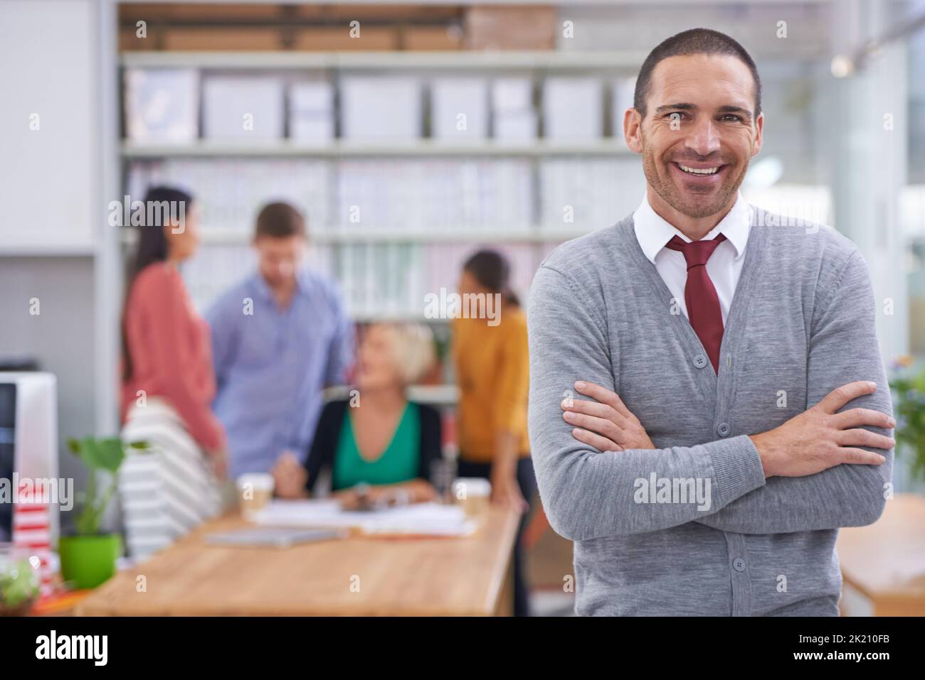 Il successo è un dato con me in carica. Un bel giovane in piedi con le braccia piegate mentre i suoi colleghi lavorano sullo sfondo. Foto Stock