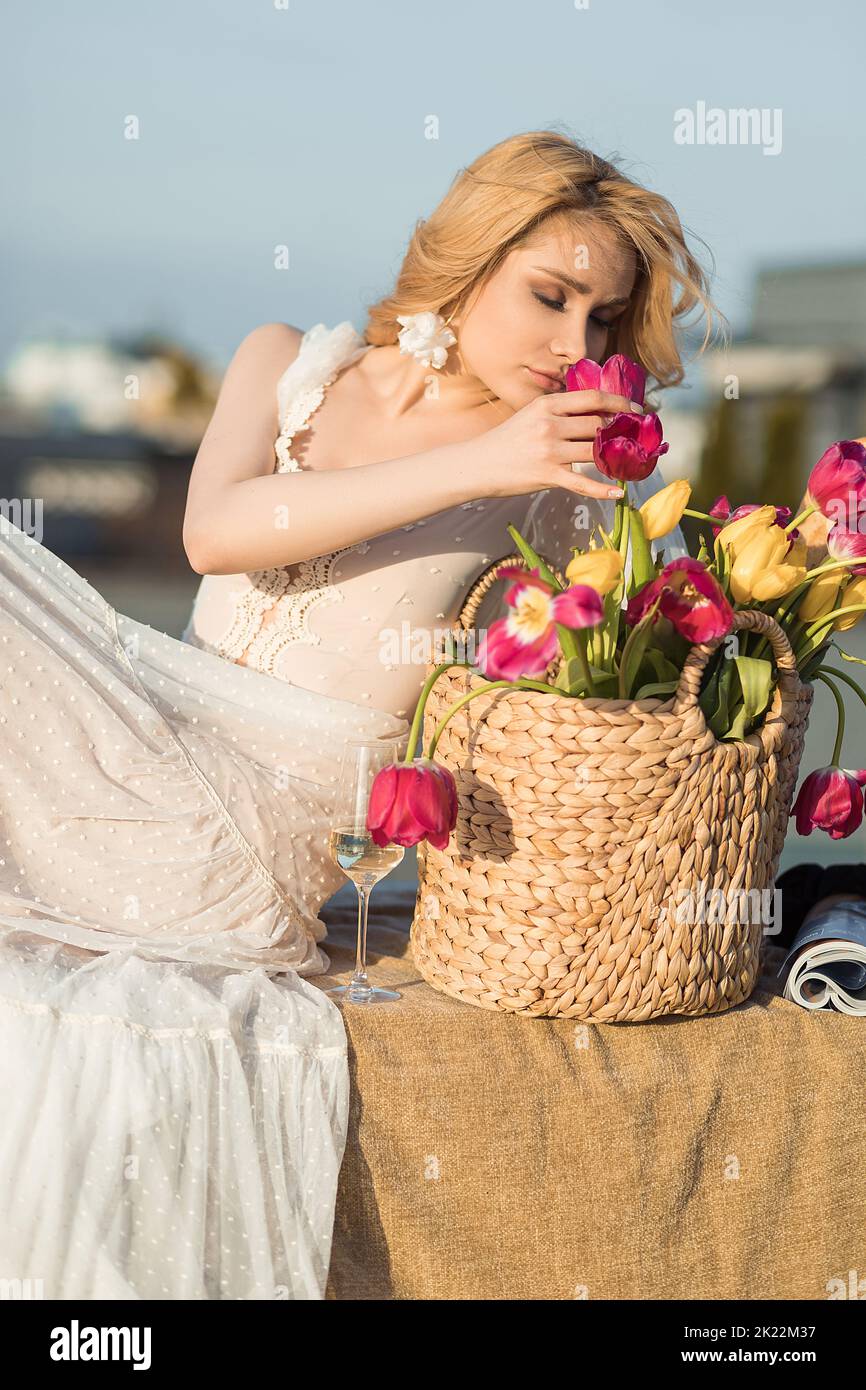 Donna di charme in abito romantico odore bouquet di tulipani rosa e giallo in cesto di vimini sulla terrazza sul tetto. Elegante signora relax con un bicchiere di champagne Foto Stock