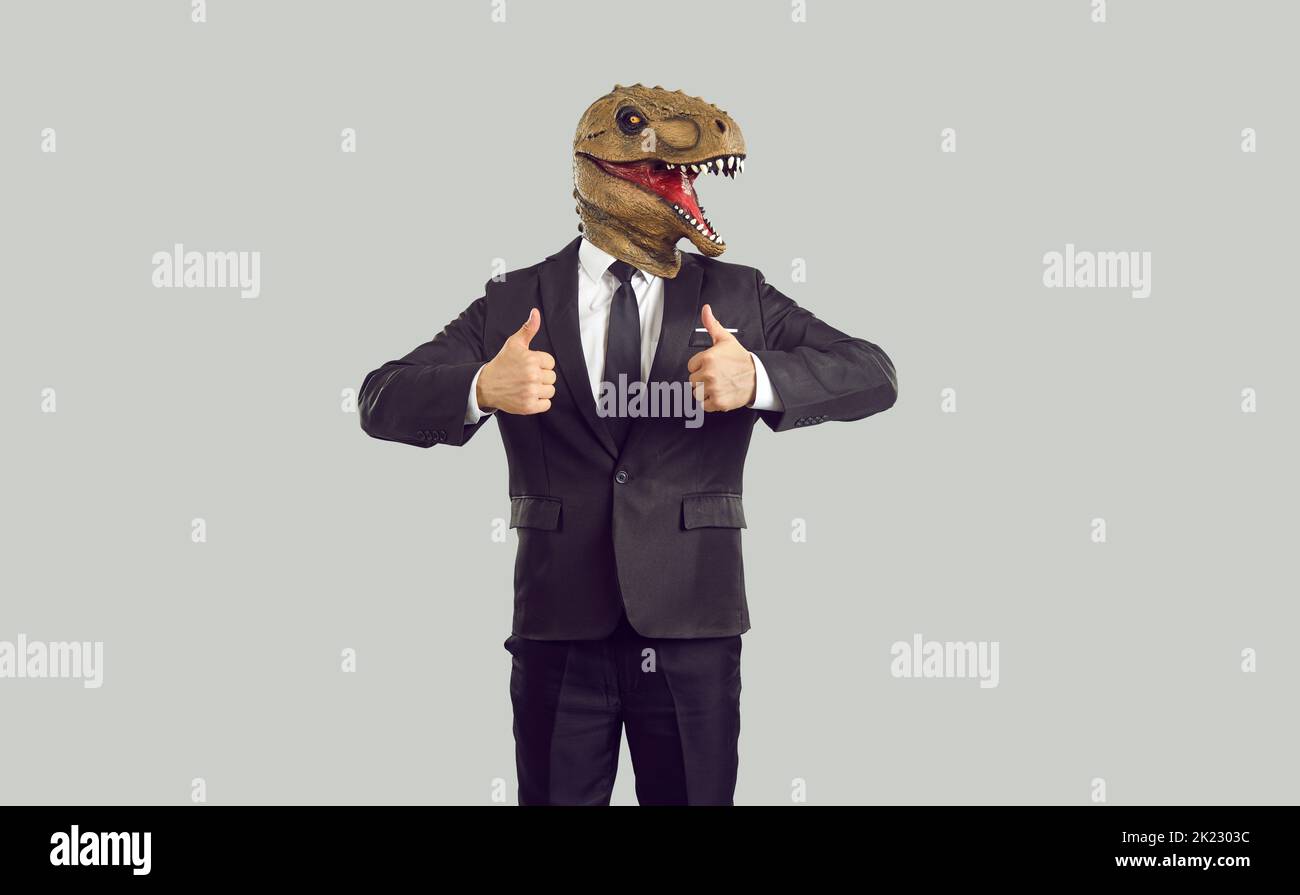 Uomo d'affari divertente in umoroso dinosauro alla moda maschera mostrando pollici su sfondo grigio. Foto Stock
