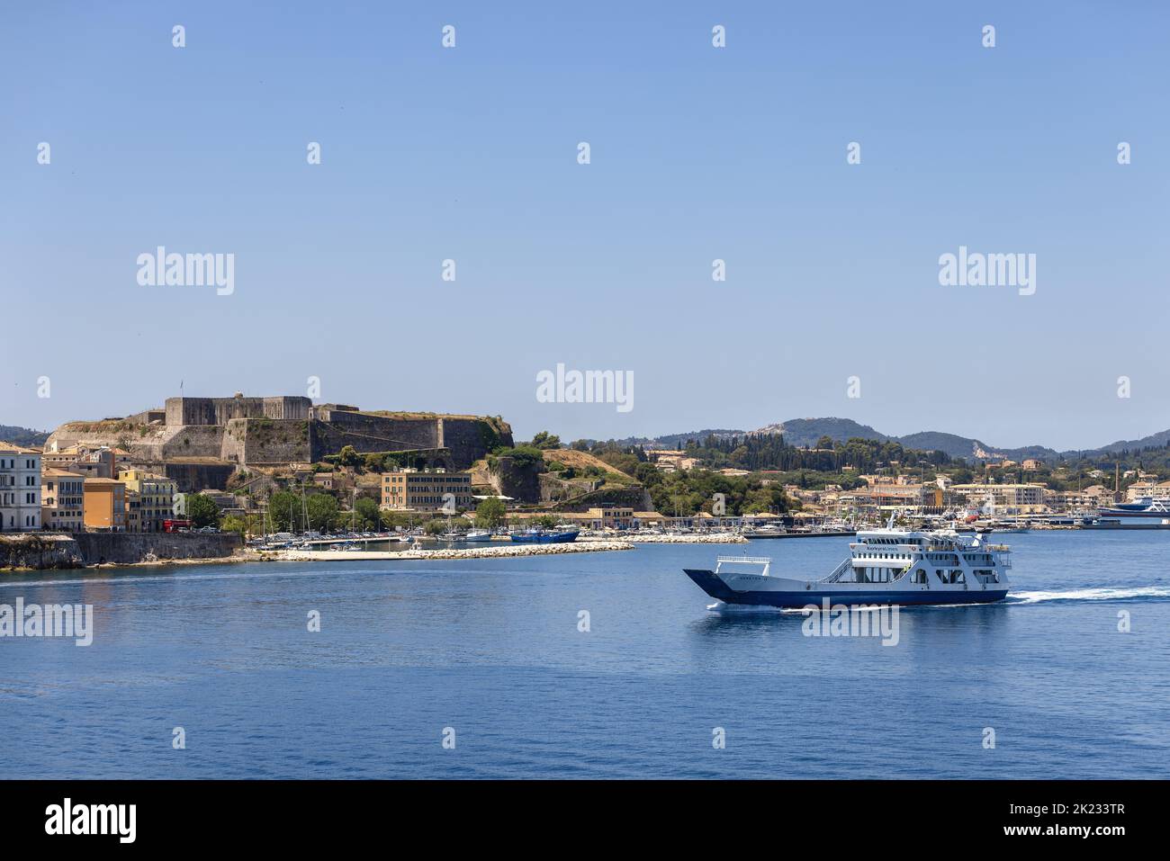 Un piccolo traghetto bianco e blu attraversa la tranquilla baia dell'isola di Corfù sullo sfondo della nuova fortezza della città, le isole ioniche, la Grecia Foto Stock