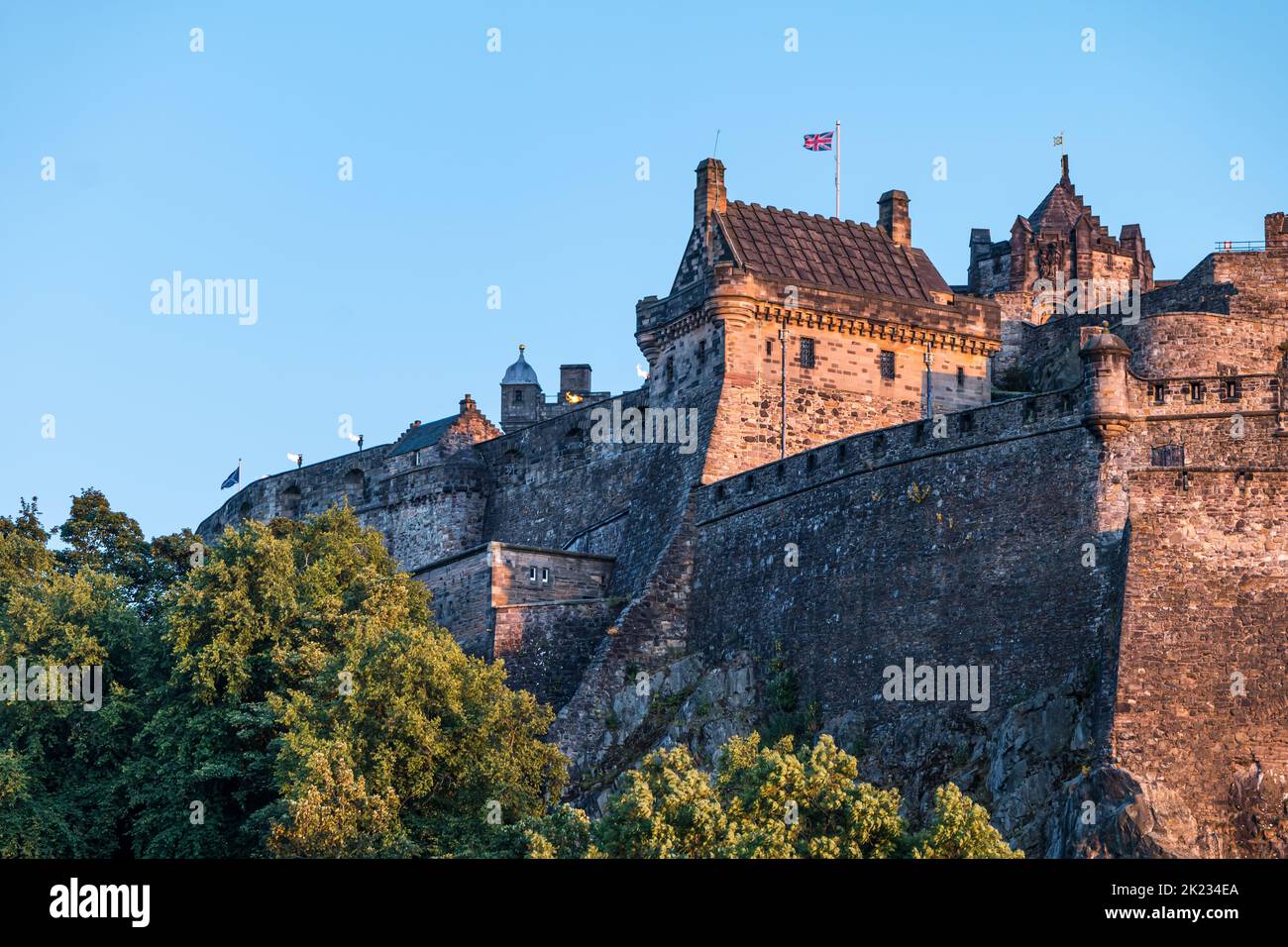 Vista del Castello di Edimburgo che batte una bandiera britannica di Union Jack dalla roccia dell'entroterra con cielo blu chiaro, Scozia, Regno Unito Foto Stock