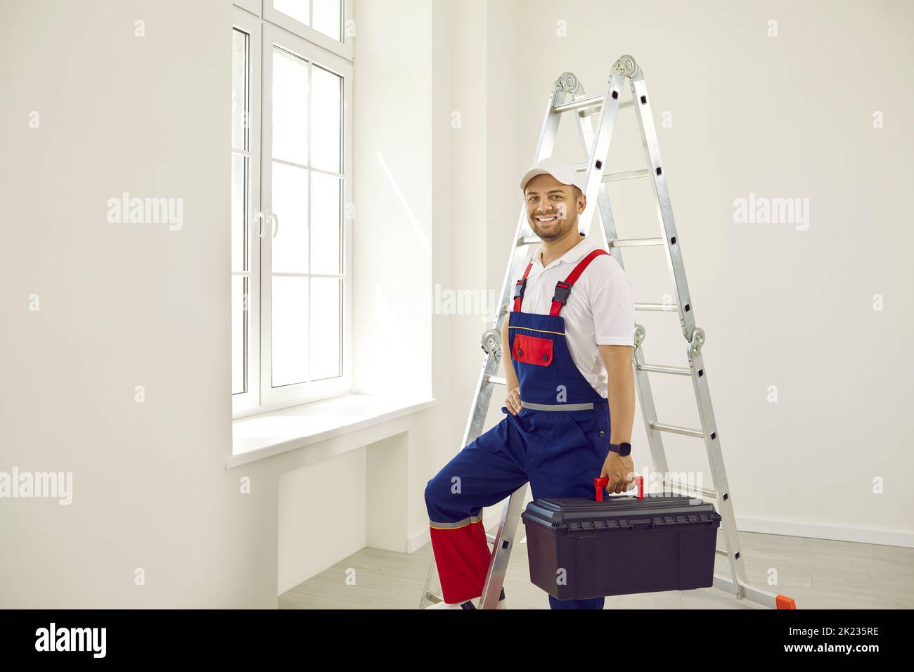 Lavoratore maschio felice in uniforme in piedi in stanza vuota, tenendo cassetta degli attrezzi e sorridendo Foto Stock