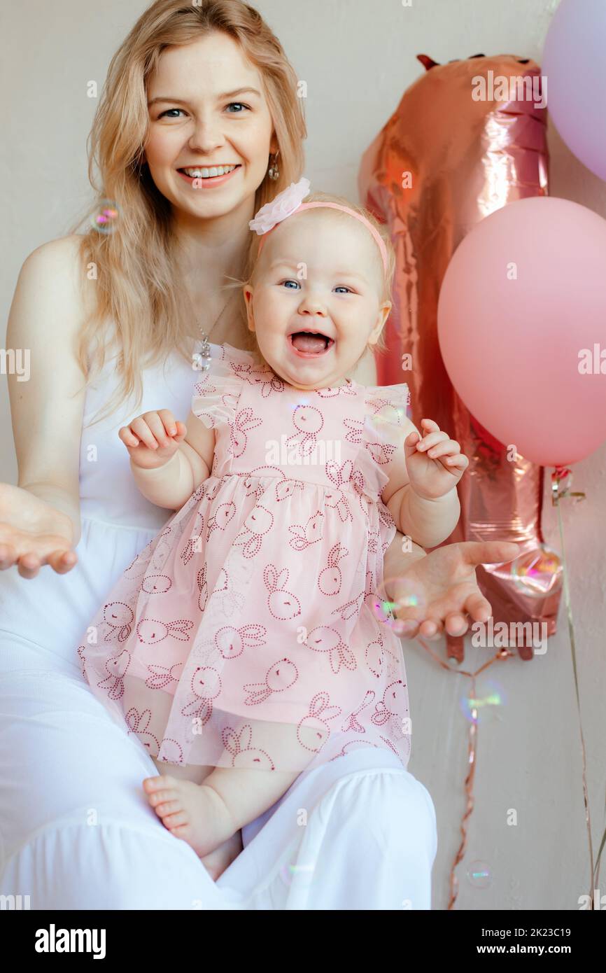 Donna bionda risata e folle, madre che tiene il bambino in vestito rosa e bandelet floreale, festeggia un anno Foto Stock