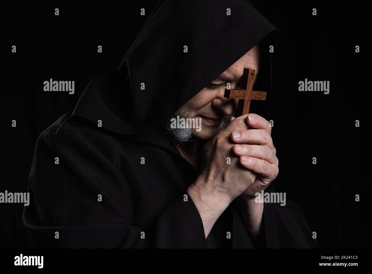 monaco anziano in cassock incappucciati che prega con crocifisso vicino alla faccia isolata su nero, immagine stock Foto Stock