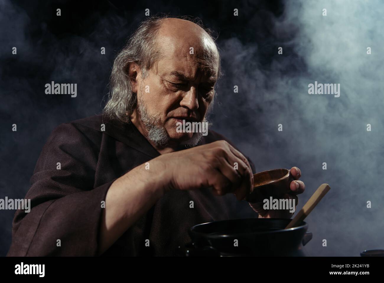 alchimista bearded che aggiunge l'ingrediente nella pentola mentre prepara la pozione su sfondo nero con fumo, immagine di scorta Foto Stock