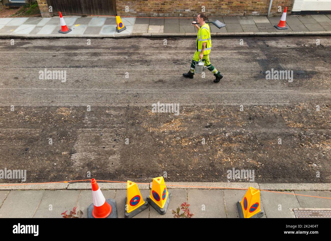 Operaio cammina per la strada con la pala sulla spalla dopo la rimozione meccanizzata della vecchia superficie stradale prima di rifare una strada residenziale a Twickenham, Greater London, UK. (132) Foto Stock