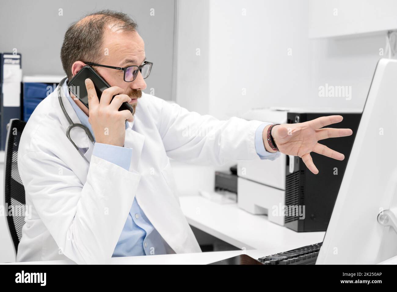 Medico di sesso maschile che punta con il dito sul computer desktop mentre parla al telefono, discutere il trattamento con il collega. Foto di alta qualità Foto Stock