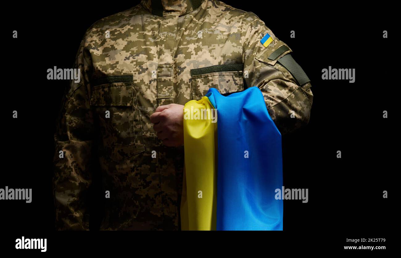Un soldato delle forze armate ucraine si trova con una bandiera blu-gialla dell'Ucraina su sfondo nero. Onorare i veterani e commemorare quelli uccisi in guerra Foto Stock