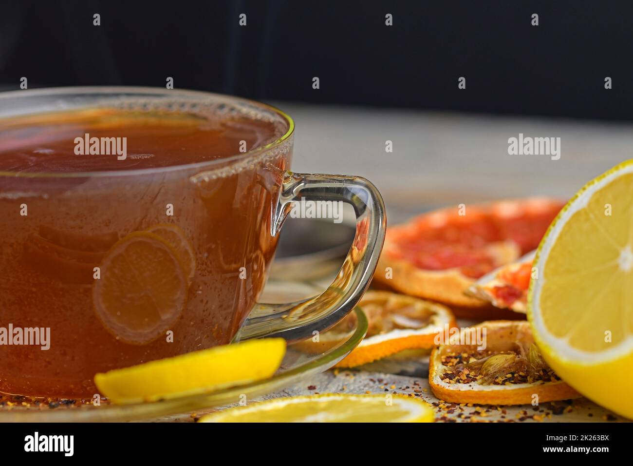 Tazza con tè caldo e vapore sul nero. Bicchiere di tè nero con bastoncini di cannella, anice stella, limone e frutta secca su fondo tavolo di legno bianco. Immagine macro Foto Stock