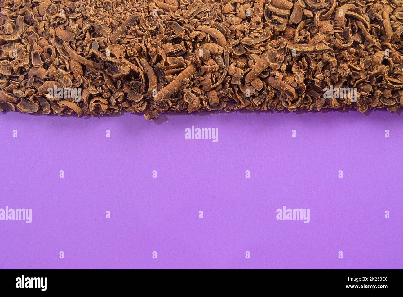 Consistenza di scaglie di cioccolato al latte. Sfondo di cioccolato macinato. Cioccolato al latte grattugiato sparso. Disposizione piatta e grande spazio di copia. Scaglie di cioccolato su sfondo lilla Foto Stock