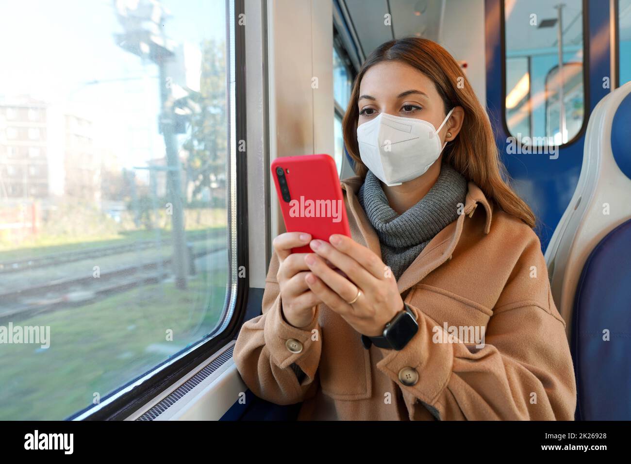 Giovane donna che indossa la maschera facciale FFP2 KN95 contro la lettura del coronavirus su smartphone che viaggia con i mezzi pubblici. Concetto di salute, protezione dai virus e sicurezza di viaggio. Foto Stock