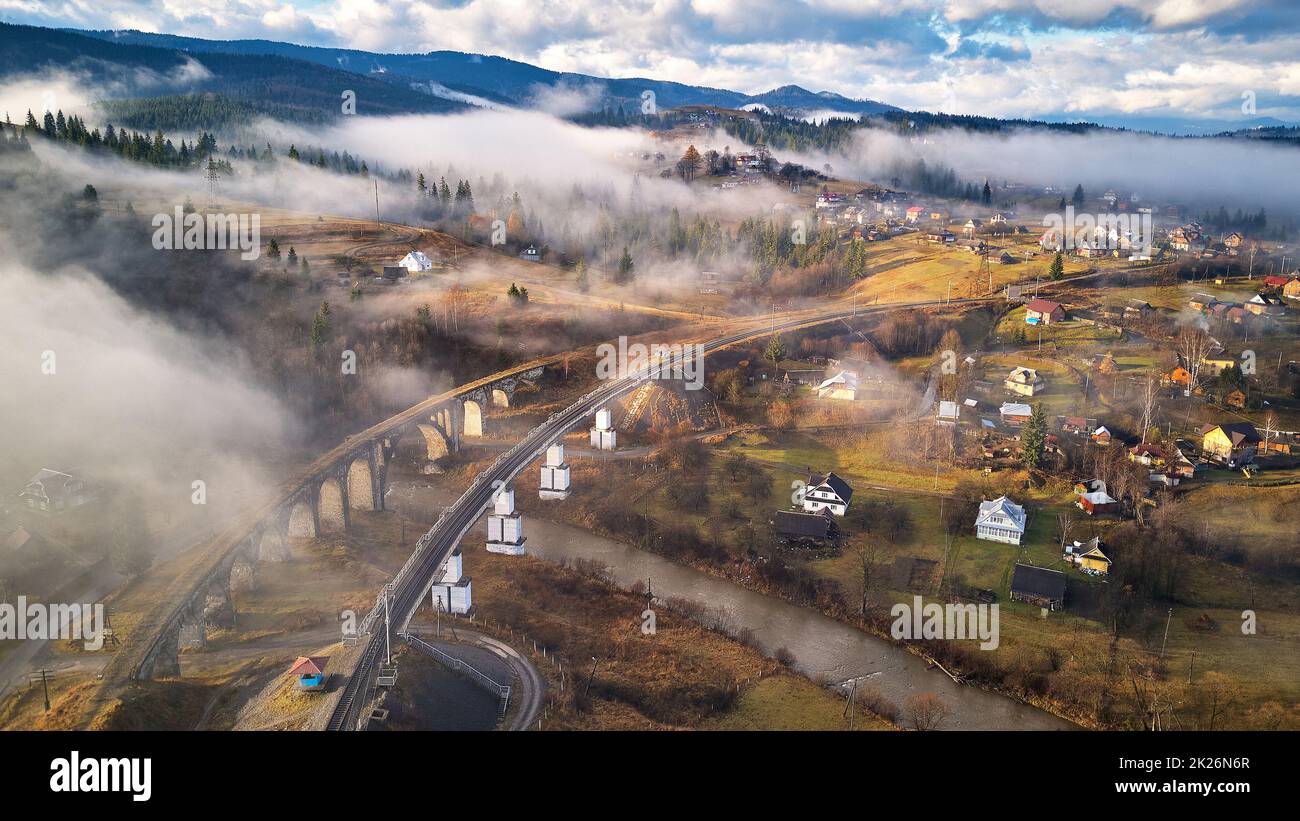 Mattina d'autunno, valle di montagna nebbiosa. Ponte ferroviario, vecchio viadotto in Vorohta. Villaggio sulle colline coperte da nuvole. Autunno paesaggio rurale Foto Stock