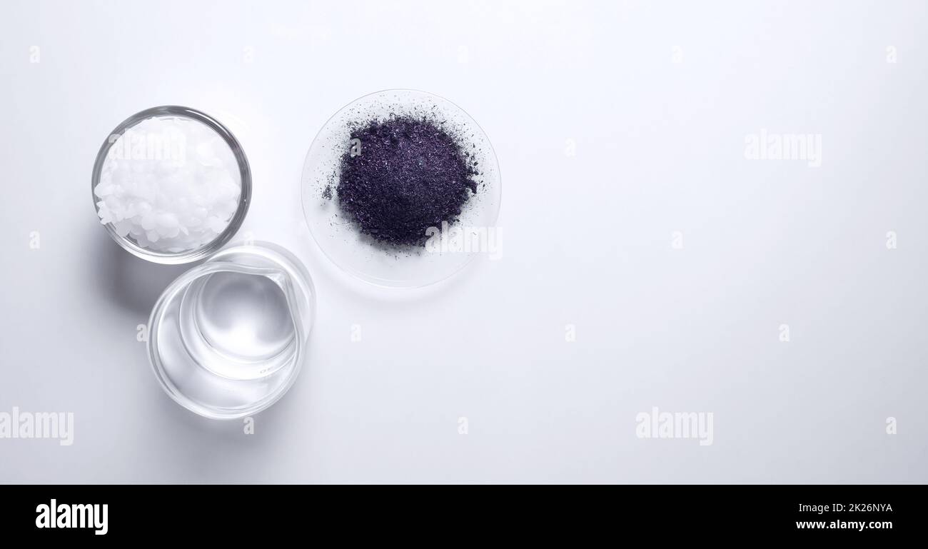 Ingrediente chimico cosmetico su tavolo bianco da laboratorio (vista dall'alto). Cera microcristallina in contenitore di vetro, KMnO4 in vetro da orologio chimico e alcool in becher. Foto Stock