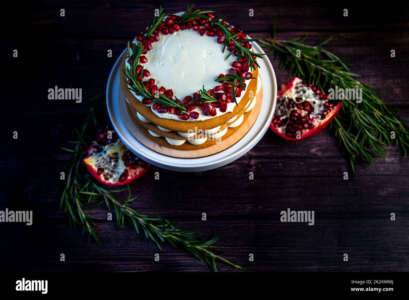 Torta con crema bianca e dolci marroni al miele si trova su un banco bianco, decorato con melograno e rosmarino, contro un albero scuro Foto Stock