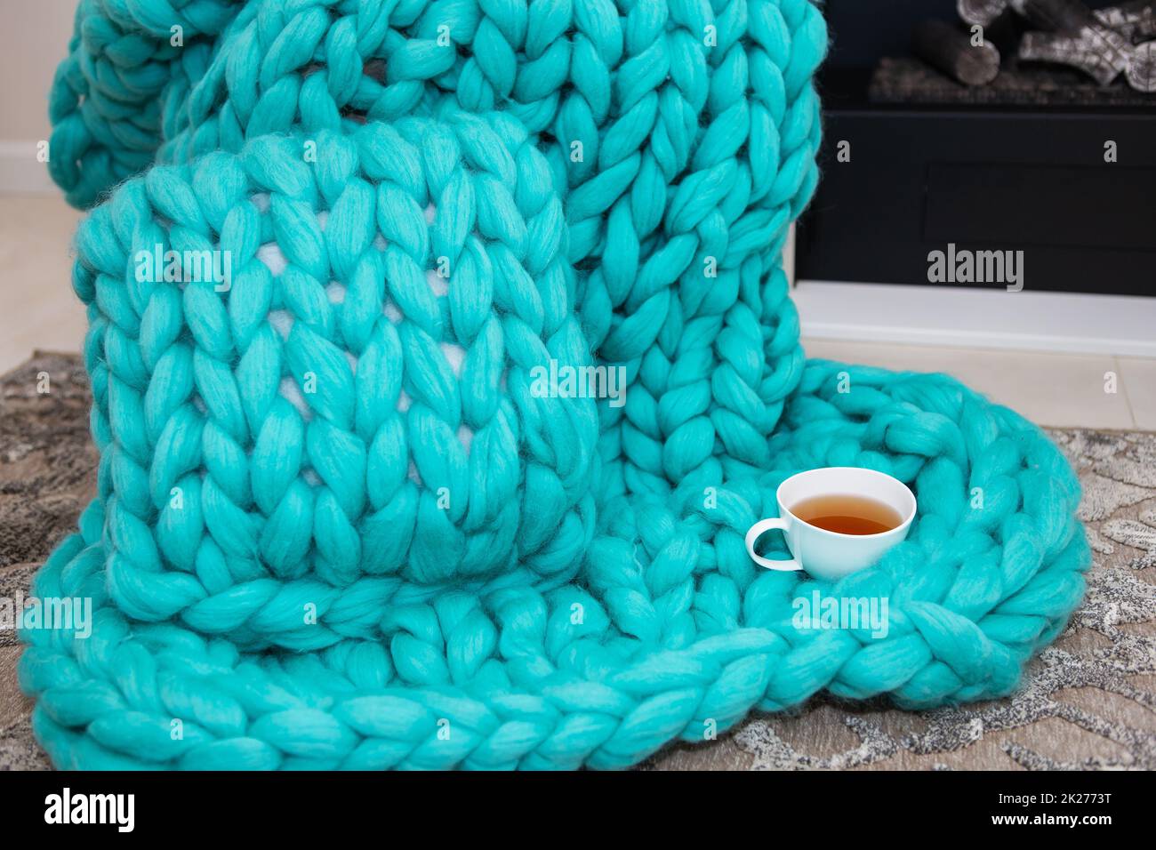 Un caldo plaid lavorato a maglia di lana merino di una bella ombra acquamarina si trova vicino al camino insieme a una tazza di tè. Il concetto di comfort. Foto Stock