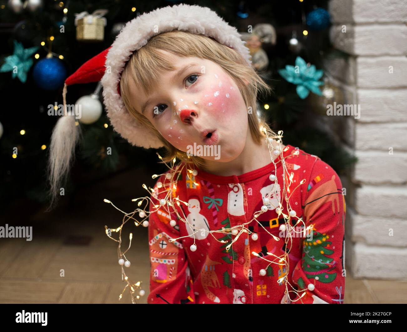 Bambino nei pigiami di Natale, cappello di Santa e un make-up del cervo dipinto sul suo volto rende divertenti volti. Natale, Capodanno, buone vacanze invernali, coccole Foto Stock