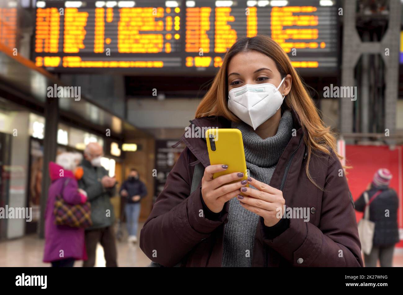 Giovane donna d'affari che indossa una maschera facciale KN95 FFP2 alla stazione ferroviaria. Giovane donna caucasica con dietro gli orari di partenza arrivi con smartphone in stazione ferroviaria. Foto Stock