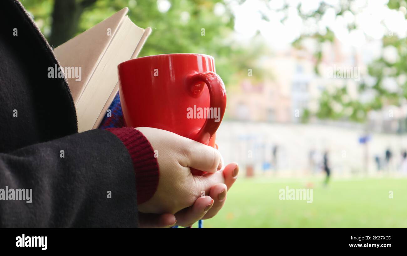 Una tazza di caffè rossa e un libro con una coperta di lana a scacchi blu o un plaid nelle mani di una donna che indossa un maglione e un cappotto nero nel parco. Tempo caldo e soleggiato. Fotografia morbida e accogliente Foto Stock