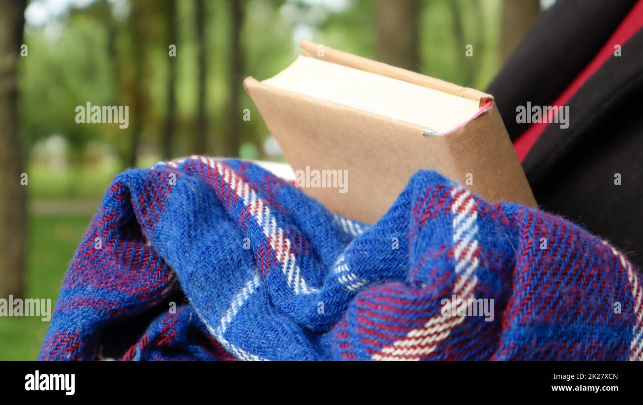 Un libro con una coperta di lana a scacchi blu o un plaid nelle mani di una donna che indossa un maglione e un cappotto nero nel parco. Tempo caldo e soleggiato. Fotografia morbida e accogliente. Primo piano Foto Stock