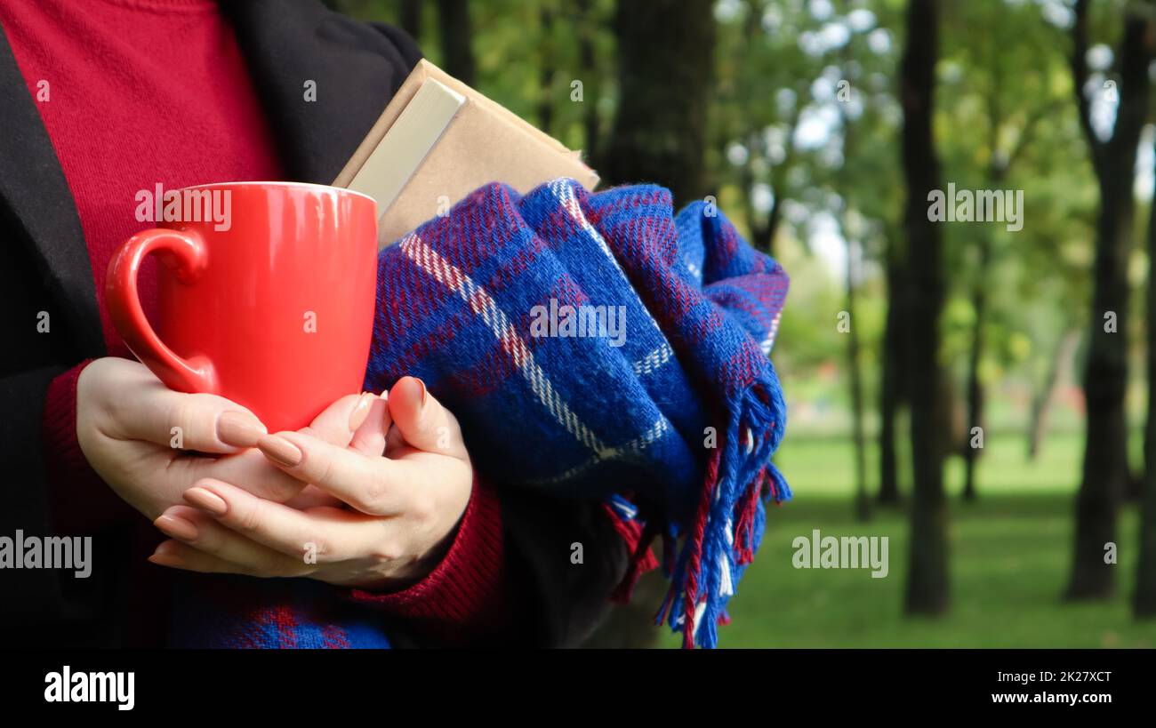 Una tazza di caffè rossa e un libro con una coperta di lana a scacchi blu o un plaid nelle mani di una donna che indossa un maglione e un cappotto nero nel parco. Tempo caldo e soleggiato. Fotografia morbida e accogliente Foto Stock