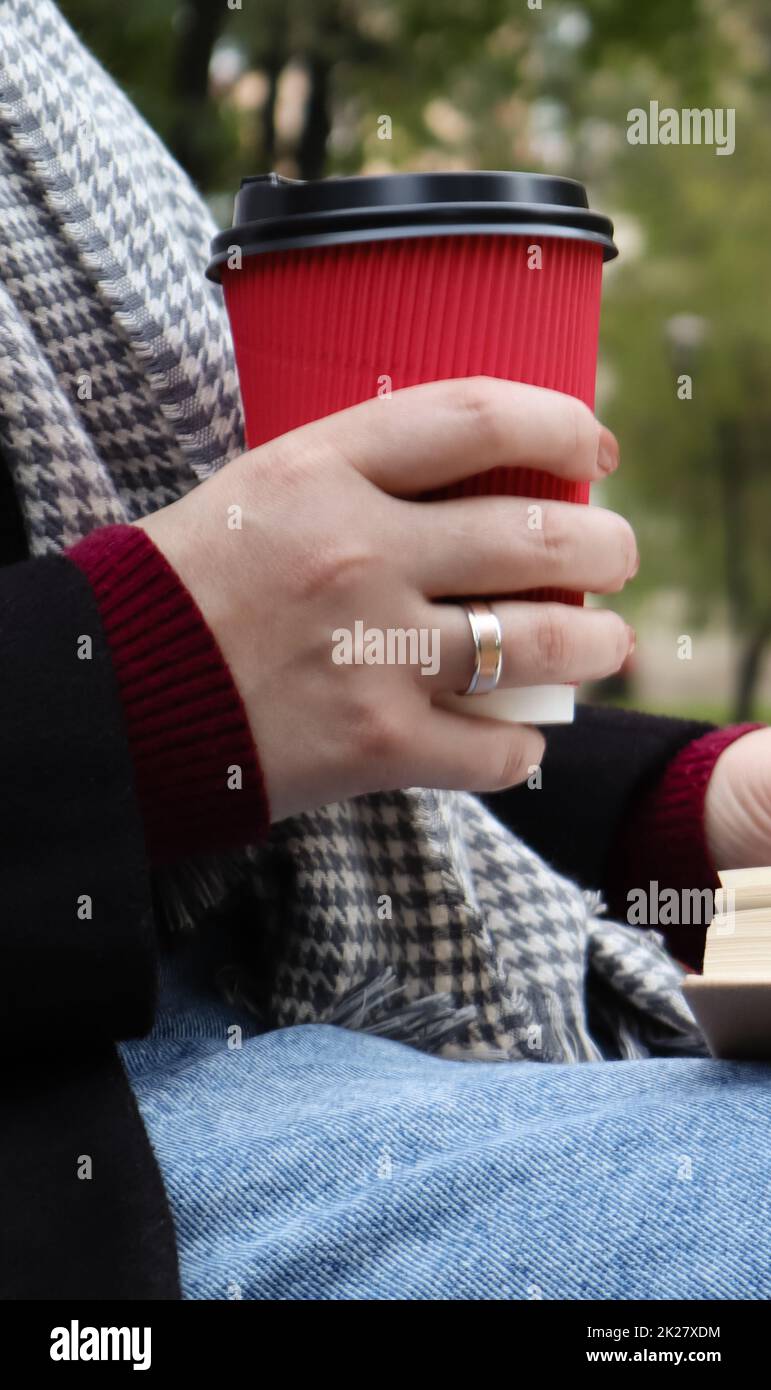 Giovane donna in jeans, cappotto e sciarpa, su una panca del parco. Una donna sta leggendo un libro e bevendo caffè o altre bevande calde all'aperto da sola. Primo piano. Il concetto di onore, studio, tempo libero e ricreazione. Foto Stock