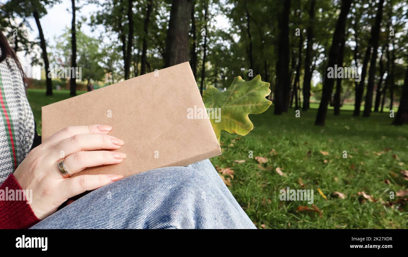 Una donna tiene un libro chiuso sdraiato in grembo con una foglia di quercia caduta primo piano in un parco in un caldo giorno di autunno soleggiato. Il concetto di relax, lettura e relax da solo. Foto Stock