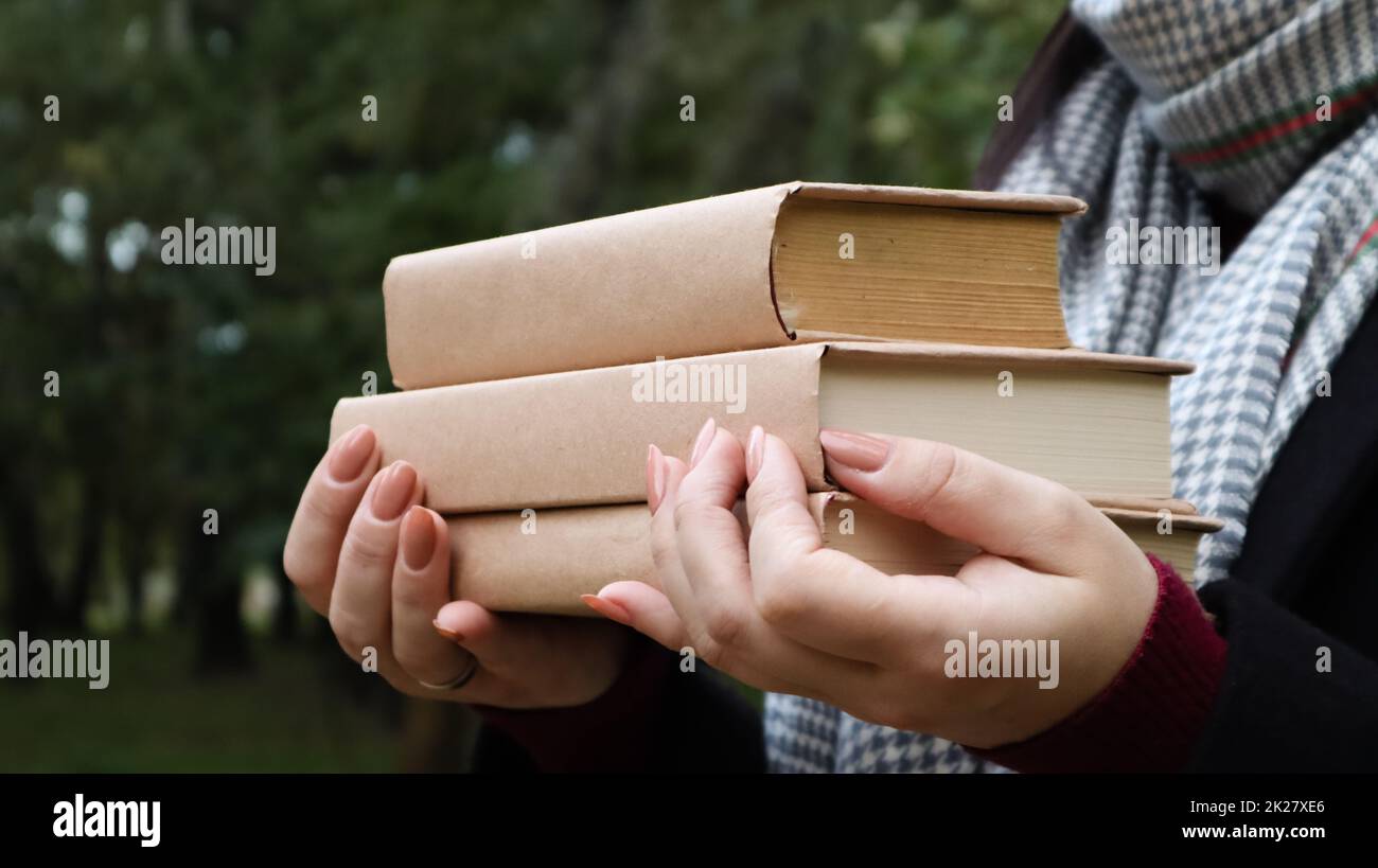 Una giovane donna in un cappotto nero e una sciarpa grigia tiene in mano una pila di libri sullo sfondo di un parco autunnale. All'aria aperta. Istruzione, mani vicine di una ragazza che studia all'università. Foto Stock