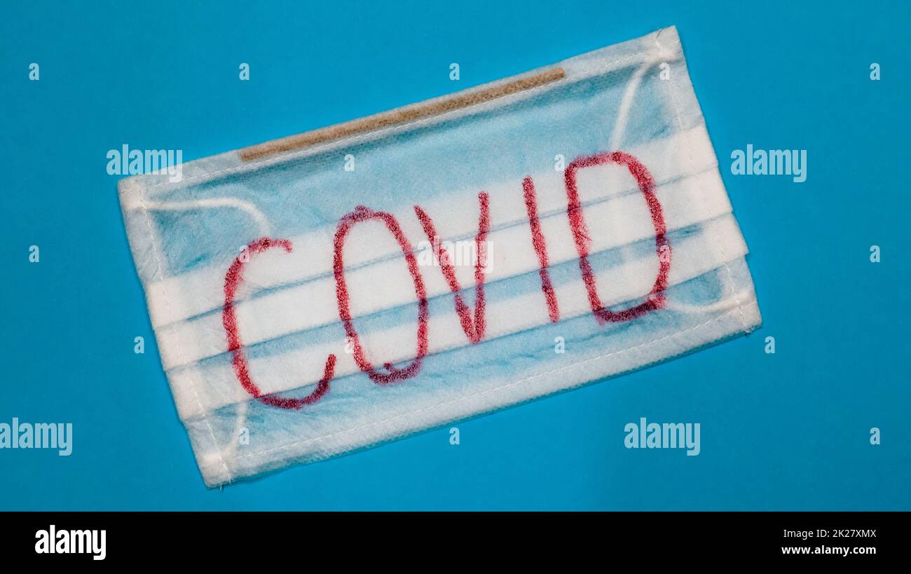 Tipica maschera chirurgica bianca a 3 strati con auricolari in gomma per coprire bocca e naso con lettere a blocco inglesi Covid su sfondo blu. Virus pericoloso e concetto di quarantena. Foto Stock