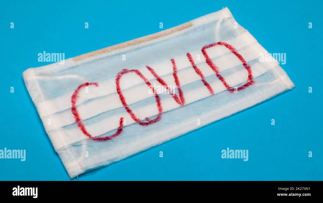 Tipica maschera chirurgica bianca a 3 strati con auricolari in gomma per coprire bocca e naso con lettere a blocco inglesi Covid su sfondo blu. Virus pericoloso e concetto di quarantena. Foto Stock