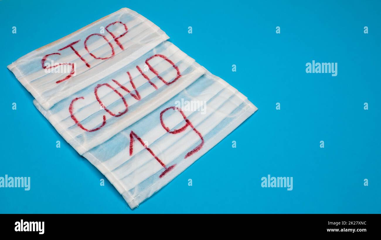 Tipica maschera chirurgica bianca a 3 veli con auricolari in gomma per coprire bocca e naso con lettere inglesi Stop Covid-19 su sfondo blu. Virus pericoloso e concetto di quarantena. Spazio di copia Foto Stock