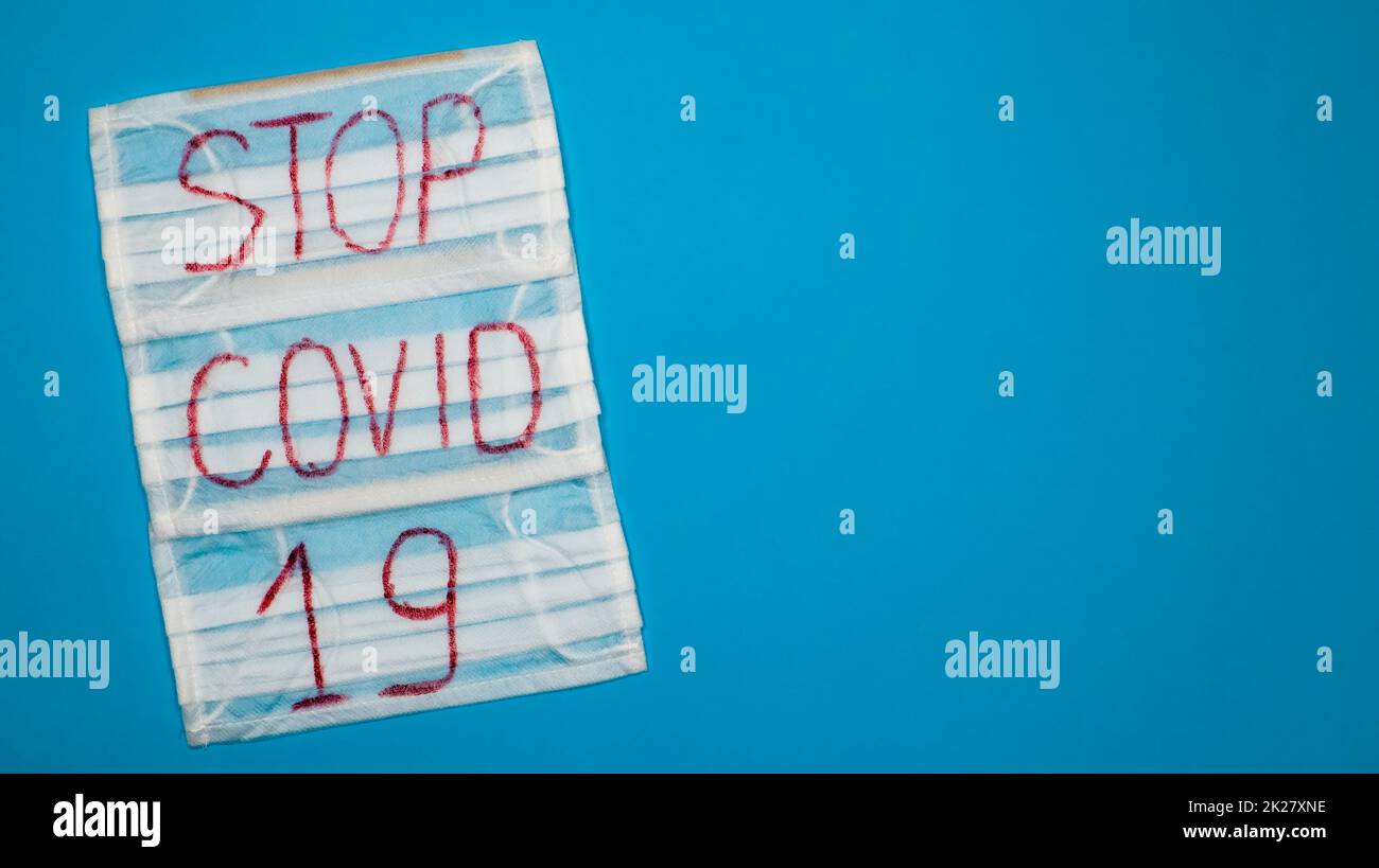 Tipica maschera chirurgica bianca a 3 veli con auricolari in gomma per coprire bocca e naso con lettere inglesi Stop Covid-19 su sfondo blu. Virus pericoloso e concetto di quarantena. Spazio di copia Foto Stock