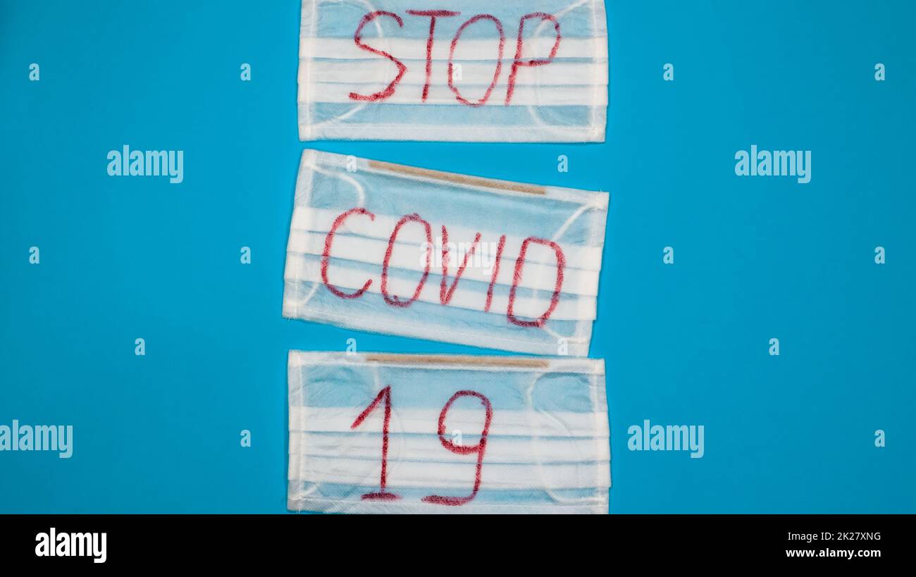 Tipica maschera chirurgica bianca a 3 veli con auricolari in gomma per coprire bocca e naso con lettere inglesi Stop Covid-19 su sfondo blu. Virus pericoloso e concetto di quarantena. Spazio di copia Foto Stock