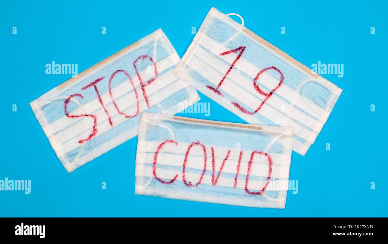 Tipica maschera chirurgica bianca a 3 veli con auricolari in gomma per coprire bocca e naso con lettere inglesi Stop Covid-19 su sfondo blu. Virus pericoloso e concetto di quarantena Foto Stock