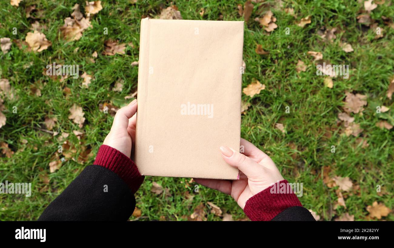 Un libro chiuso in una copertina di carta artigianale in mani femminili con erba verde e foglie gialle cadute sullo sfondo. Piano di giacitura, vista dall'alto. Modello, layout. Spazio di copia. Foto Stock