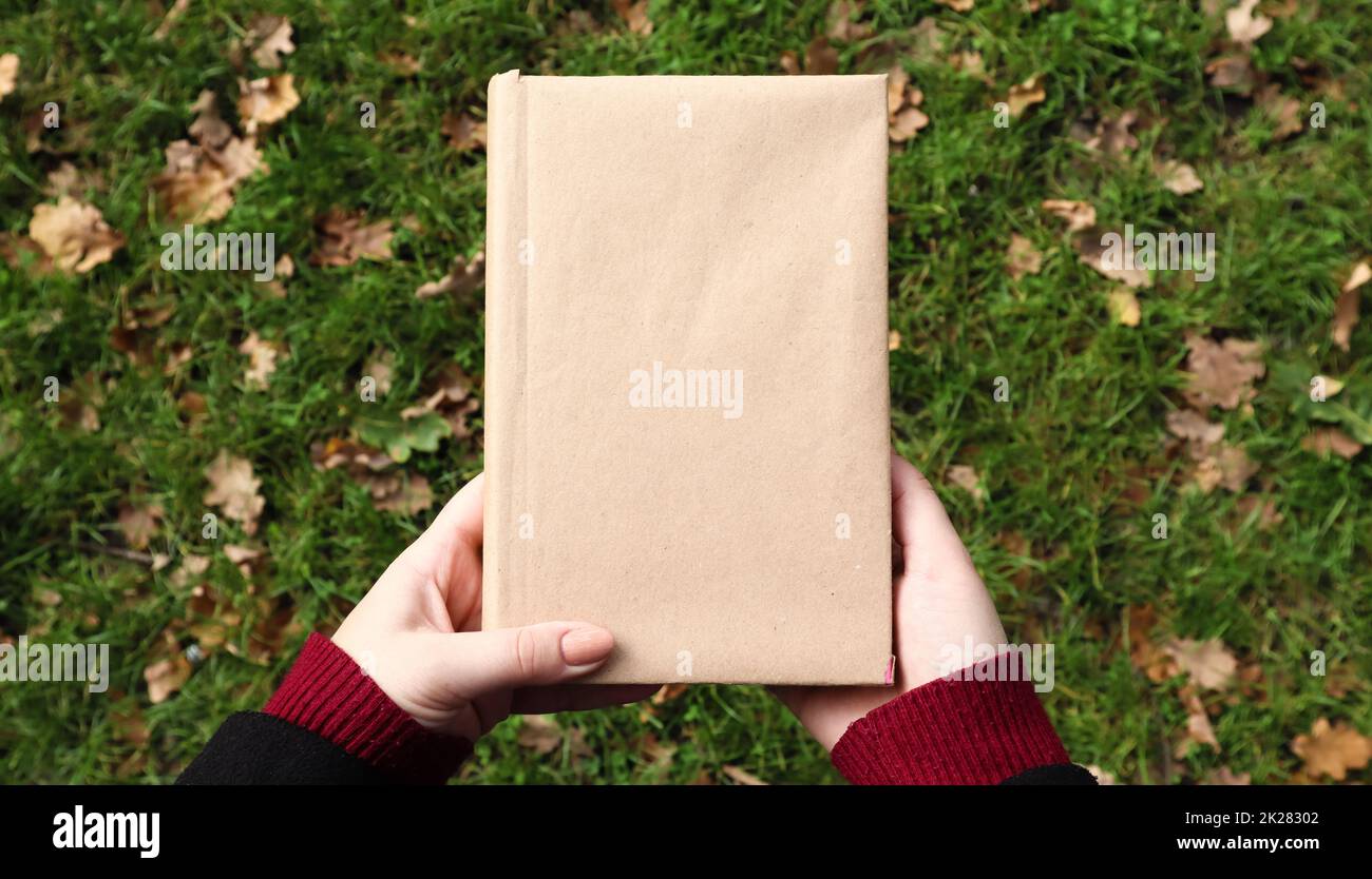 Un libro chiuso in una copertina di carta artigianale in mani femminili con erba verde e foglie gialle cadute sullo sfondo. Piano di giacitura, vista dall'alto. Modello, layout. Spazio di copia. Foto Stock