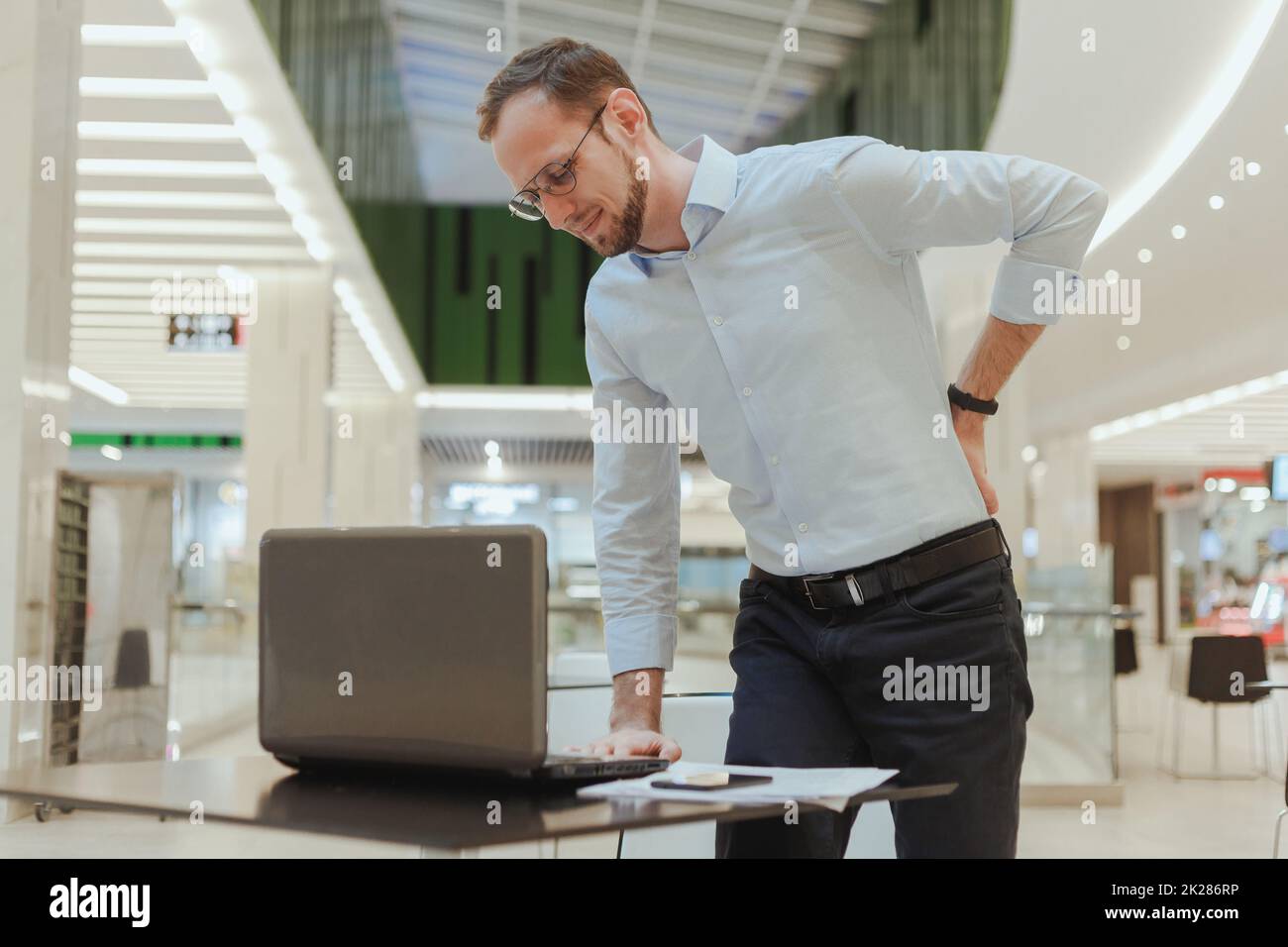 L'uomo tiene la schiena, soffre di mal di schiena, si trova vicino a un desktop con un computer. Lavoratore sedentario dell'ufficio di lavoro Foto Stock