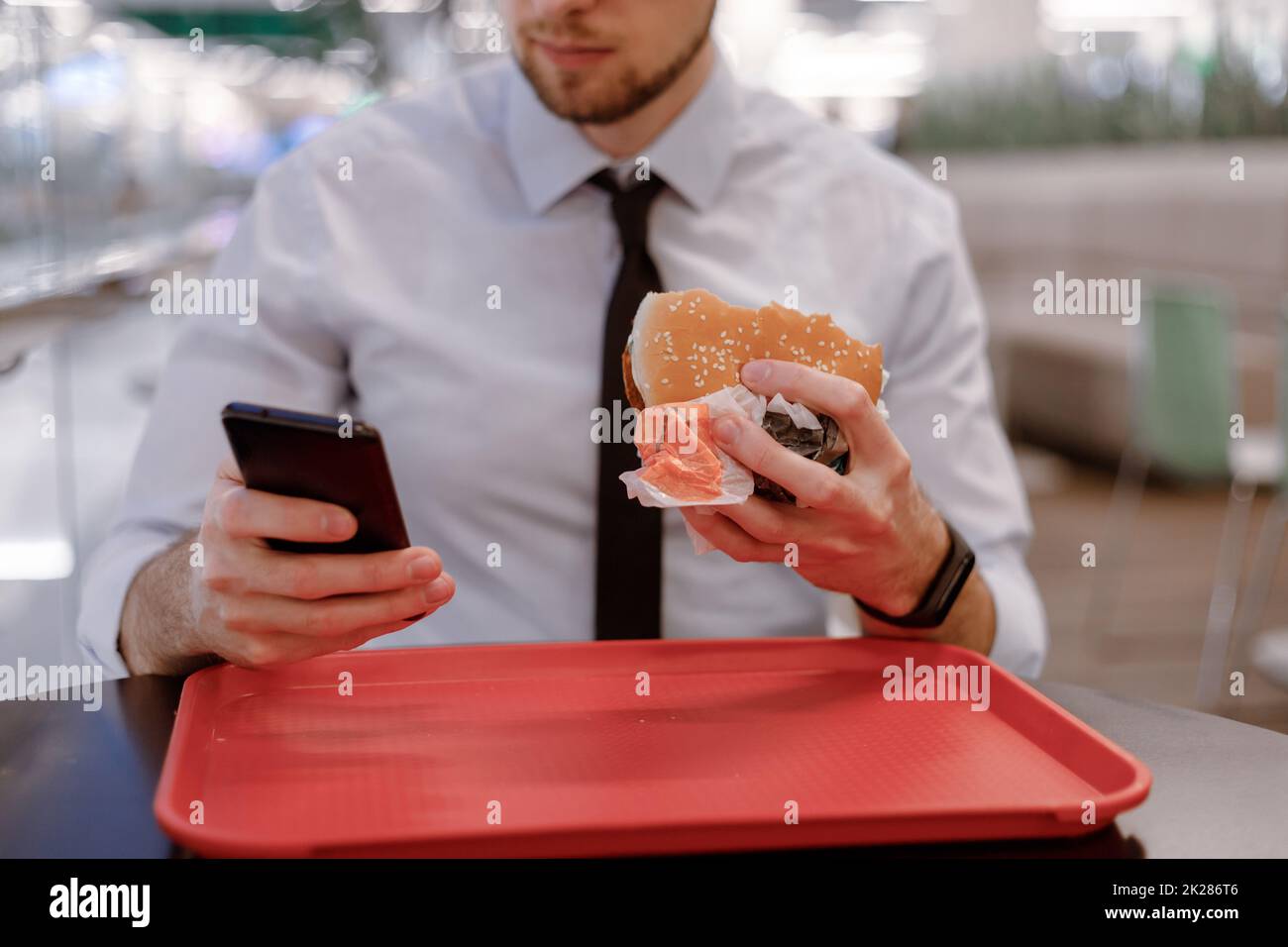 Un impiegato che si snacking a hamburger nel centro commerciale, navigando in Internet in un cellulare mentre mangia Foto Stock