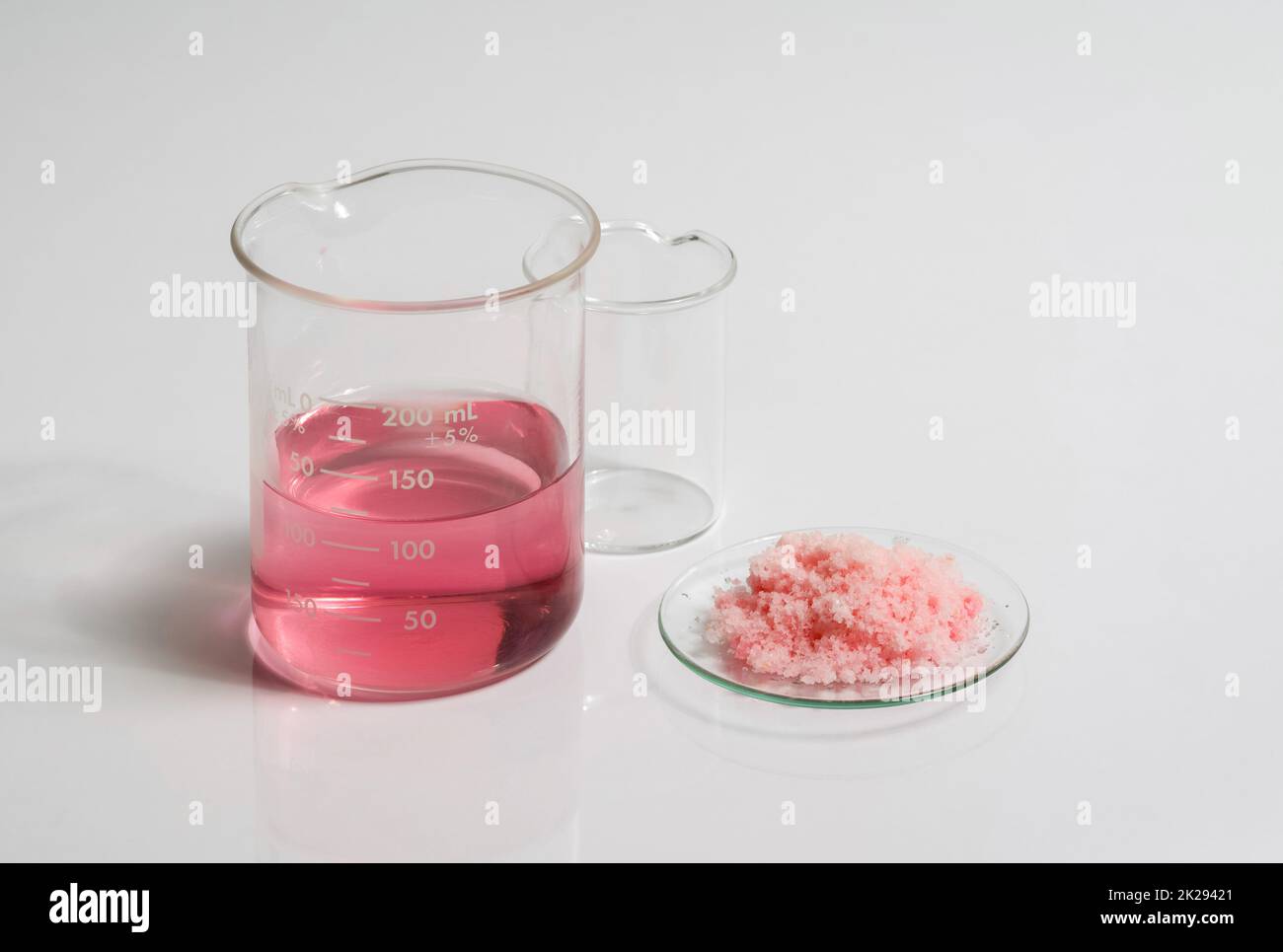 Primo piano della chimica inorganica sulla tabella bianca del laboratorio. Prodotti chimici in scaglie rosa in vetro Chemical Watch posizionare accanto alla raspina liquida EPOSSIDICA rossa nel becher. Foto Stock
