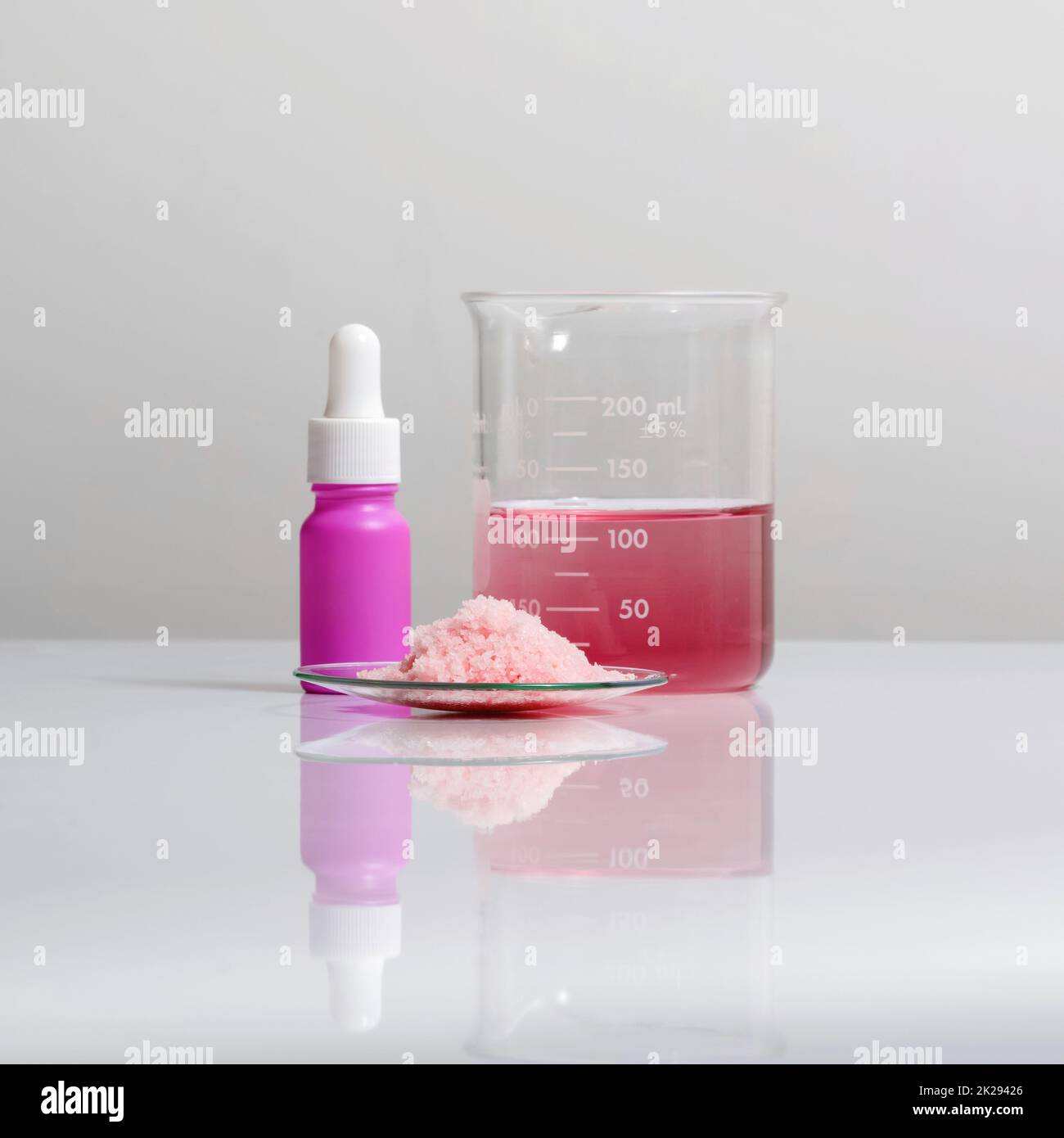 Prodotti chimici per la cura della bellezza su un tavolo da laboratorio bianco. Prodotti chimici a scaglie rosa in vetro di controllo chimico posizionare accanto al flacone di siero con contagocce e rasin liquido EPOSSIDICO rosso nel becher. Foto Stock