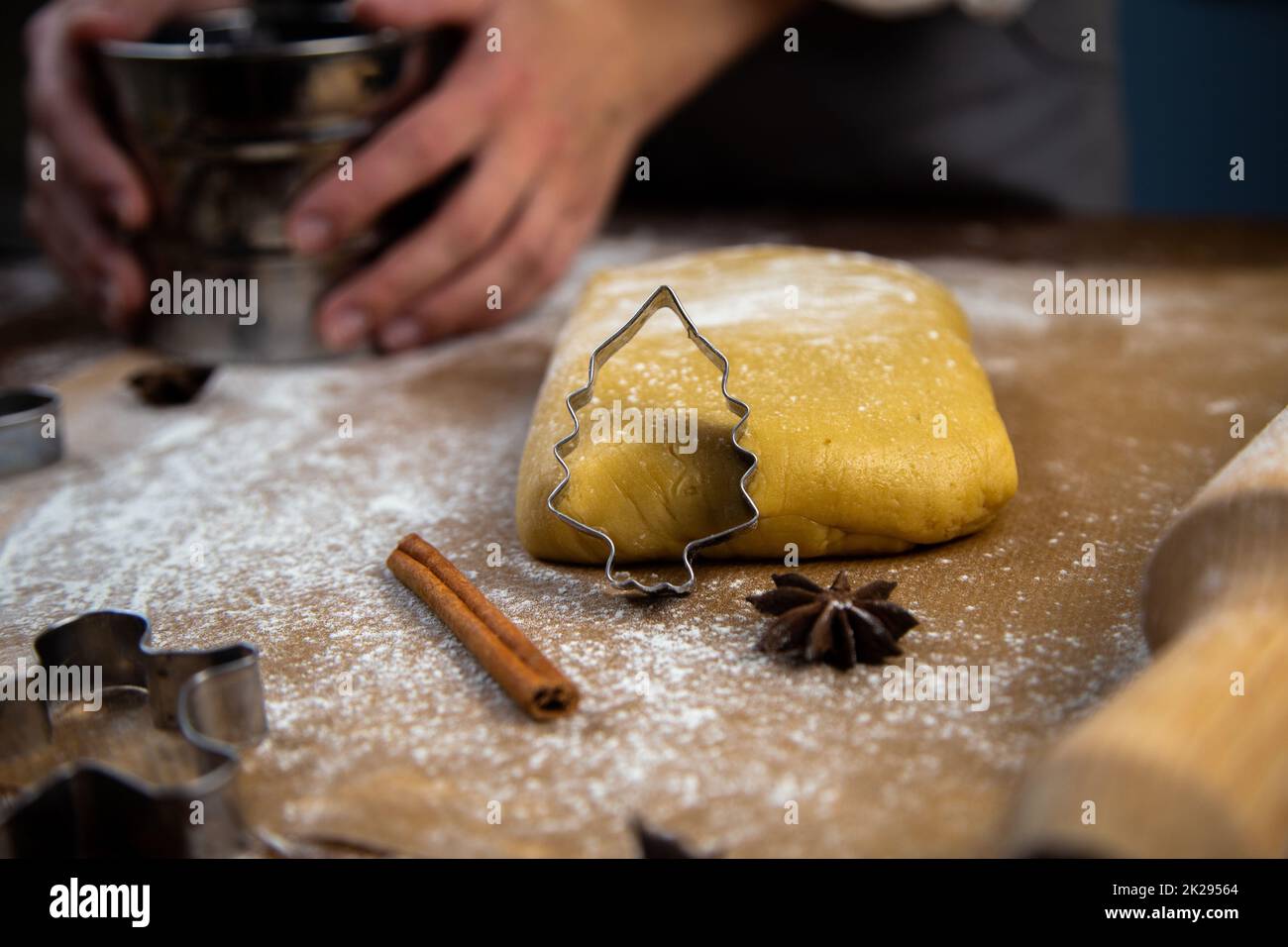Un taglialegna a forma di albero di Natale è appoggiato contro l'impasto, una foto in chiave scura, altre muffe sono visibili, una spilla, un setaccio di ferro per la farina, che è tenuto nelle mani di un pasticciere. Foto Stock