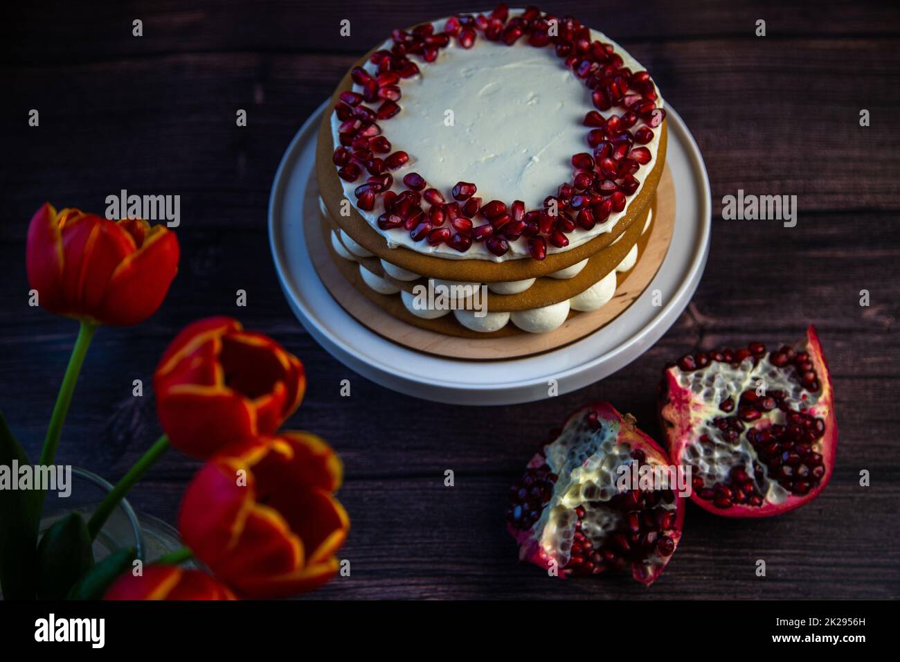 Su fondo scuro, accanto a tulipani e melograni, sorge una torta a tre strati composta da torte e crema bianca, decorata con melograno in cima a cerchio. Foto Stock