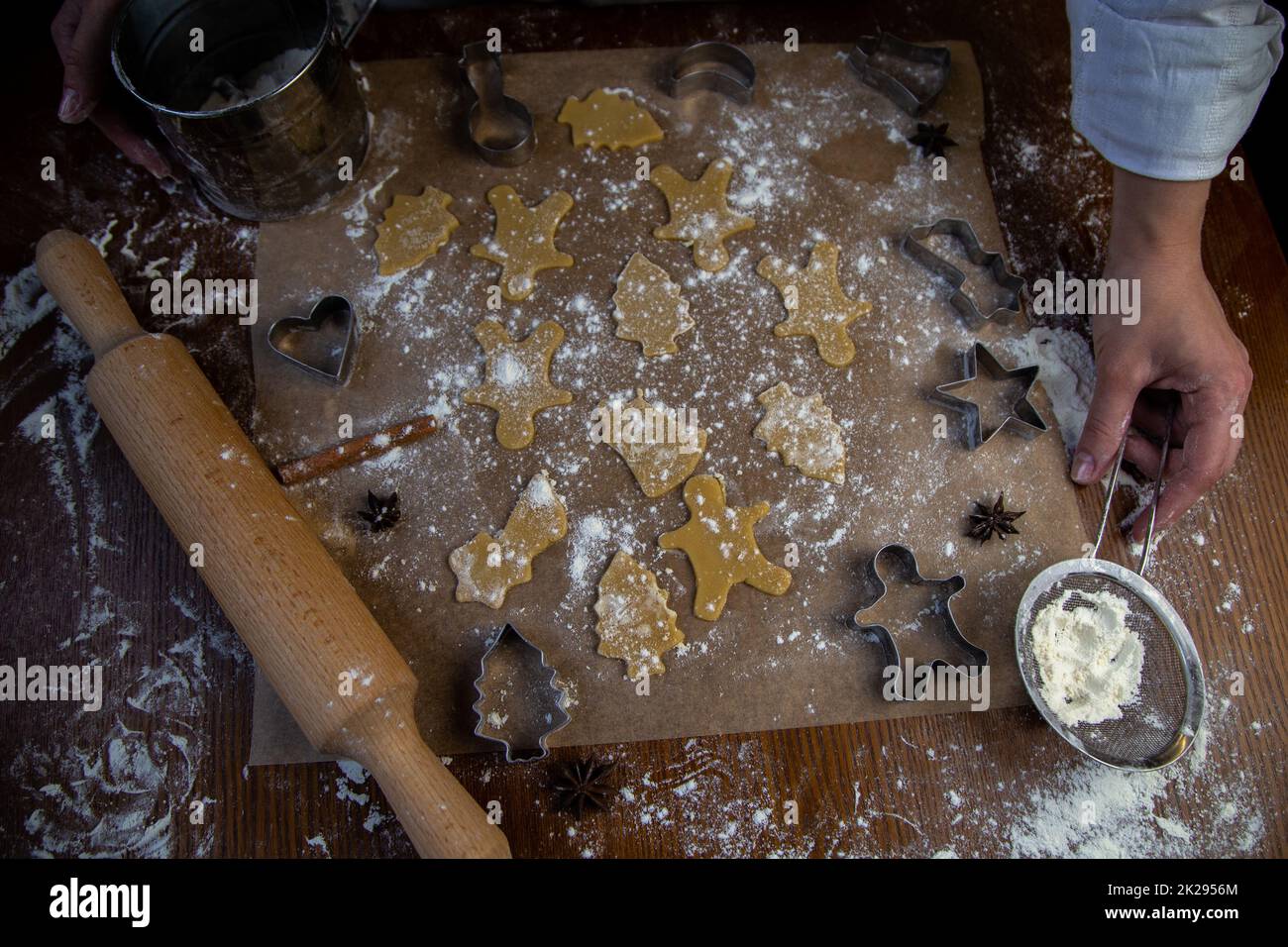 Sulla pergamena da forno sono presenti biscotti pronti di varie forme, un matterello, taglialini e un setaccio con farina sono nelle vicinanze, la farina è sparsa ovunque, le mani del pasticciere sono visibili. Foto Stock