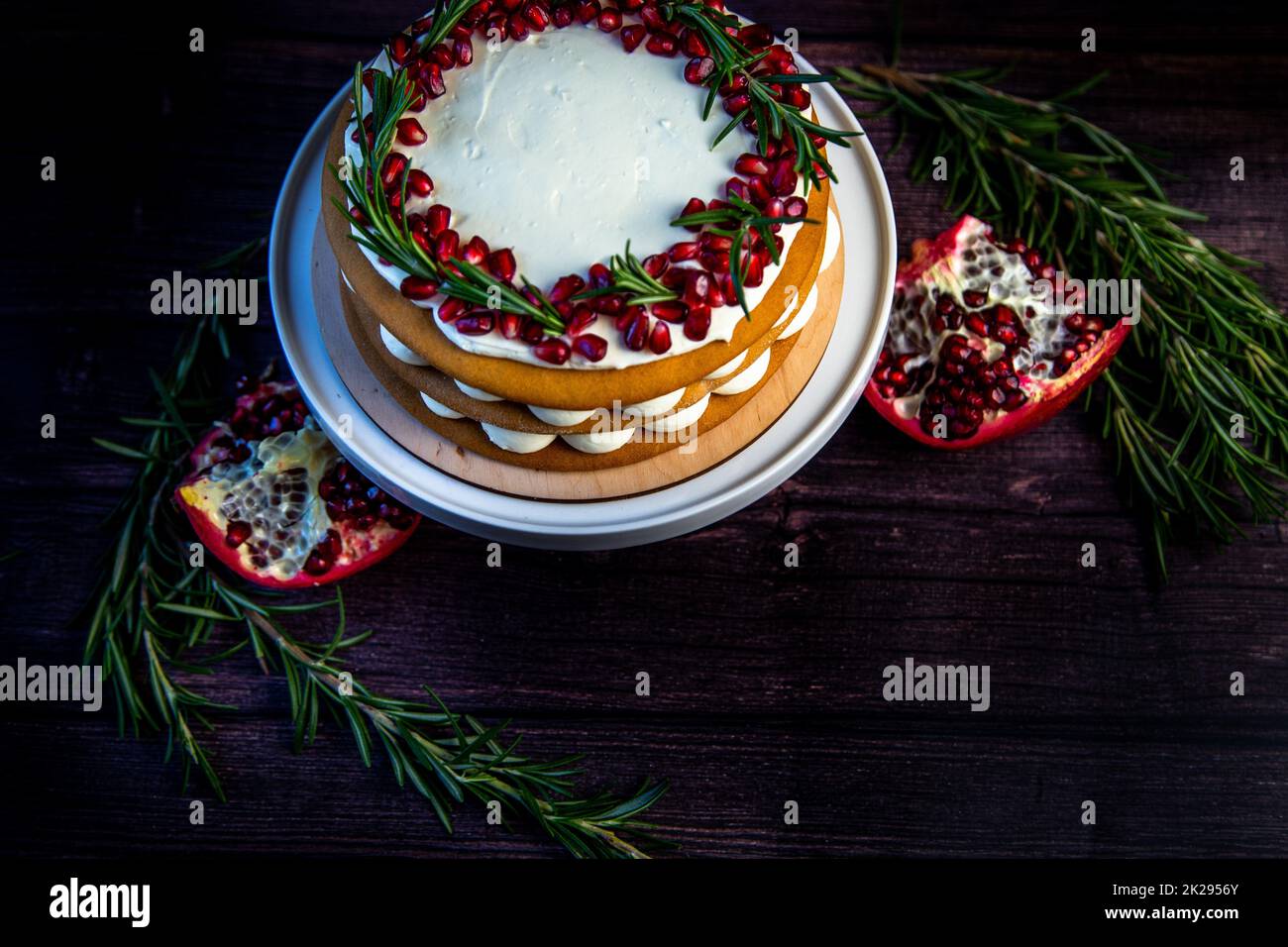 Torta con crema bianca e dolci marroni al miele si trova su un banco bianco, decorato con melograno e rosmarino, contro un albero scuro Foto Stock