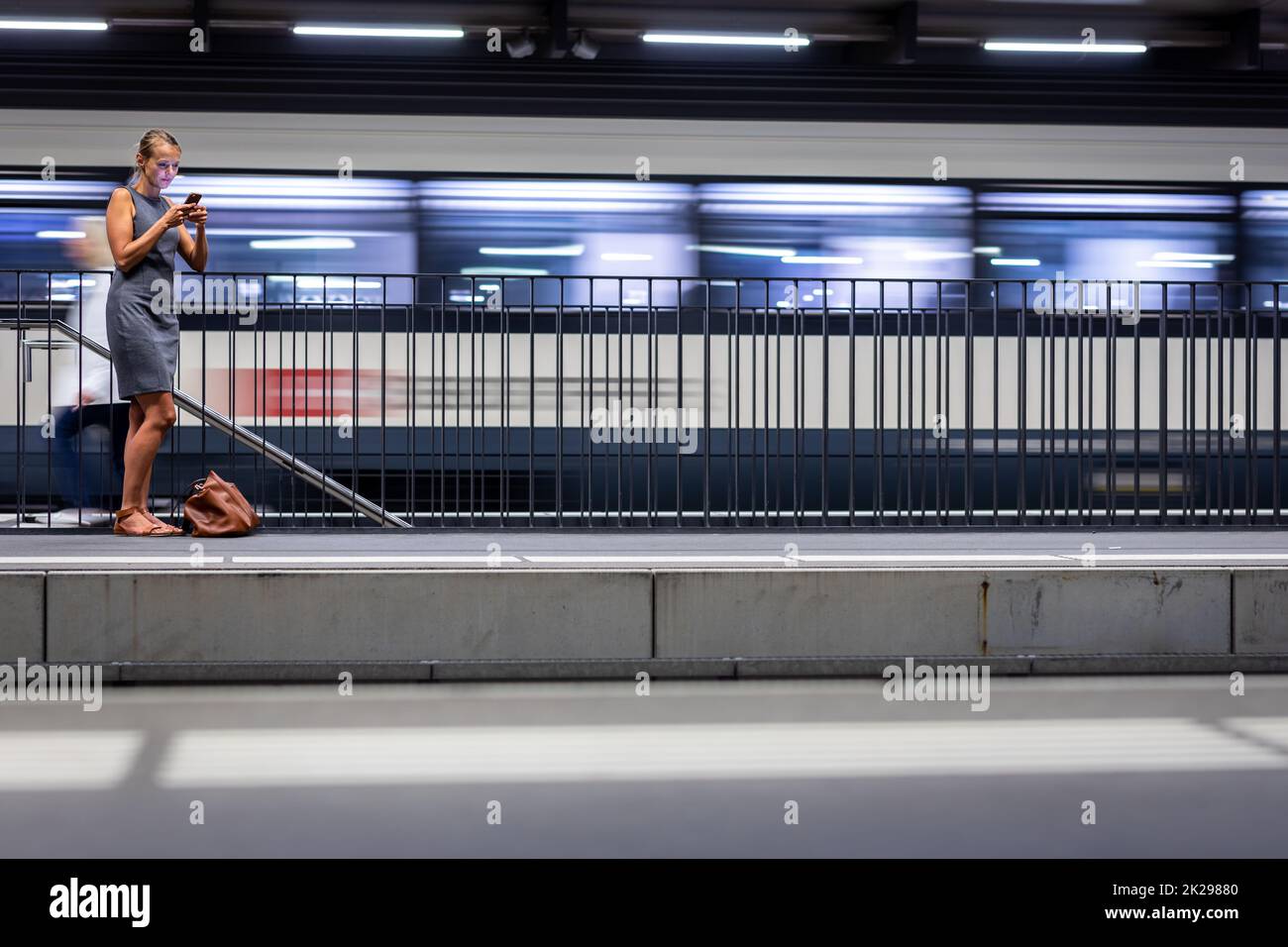 Piuttosto, giovani donne commuter in attesa del suo treno quotidiano in una moderna stazione ferroviaria, utilizzando il suo telefono cellulare mentre si è in attesa (toni di colore immagine) Foto Stock